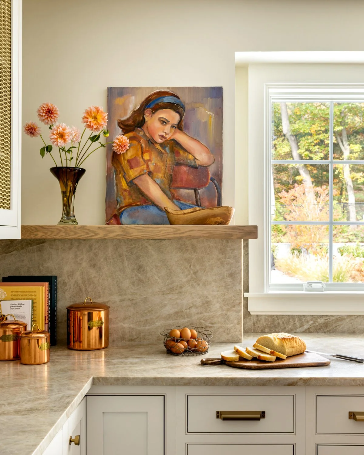 Kitchen countertop with a loaf of bread and sliced pieces, eggs in a wire basket, copper canisters, a vase with pink flowers, a painting of a woman, and a window showing autumn trees outside.