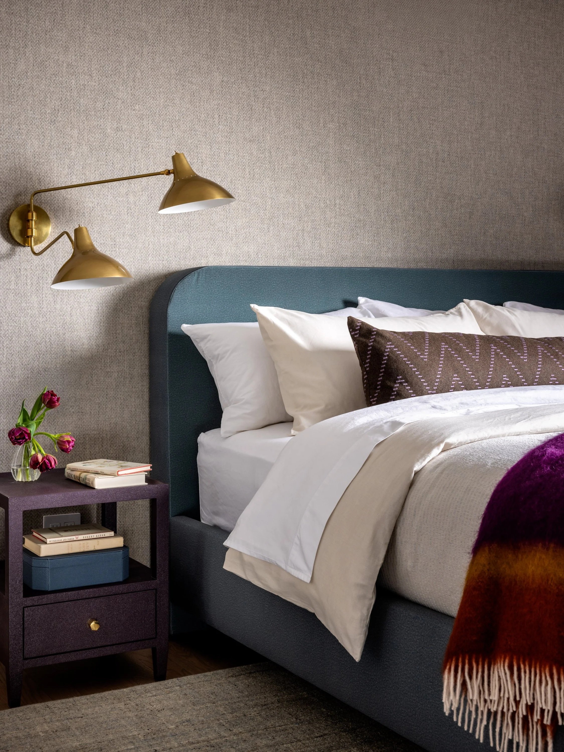 A neatly made bed with white sheets and pillows, dark brown decorative pillow, and a colorful blanket. Beside the bed is a small dark-colored nightstand with books, a vase with purple tulips, and a purple tray. Two brass wall-mounted lamps are above the bed, and textured beige wallpaper covers the wall behind the bed.