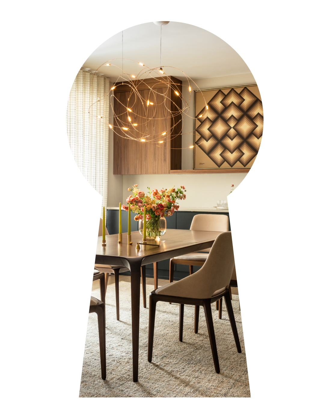 Modern dining room with a wooden table, beige chairs, floral centerpiece, green candles, and a contemporary gold chandelier. Wall art and cabinetry are visible in the background.