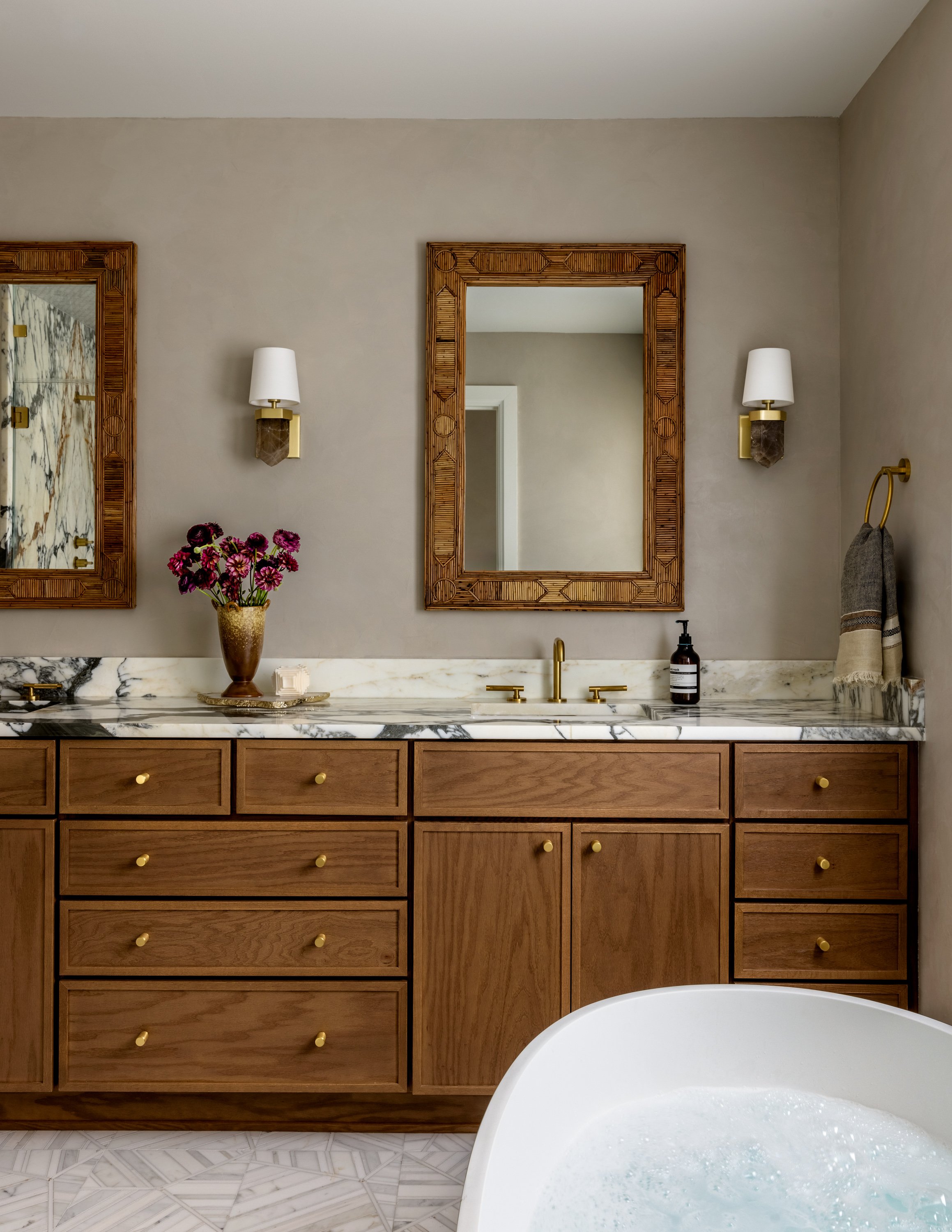 Bathroom vanity with a marble countertop, wooden cabinets, and two mirrors with wooden frames. There are wall sconces with white shades on either side of the mirrors, a vase of pink flowers, and a soap dispenser. Part of a bathtub filled with water is visible in the foreground.