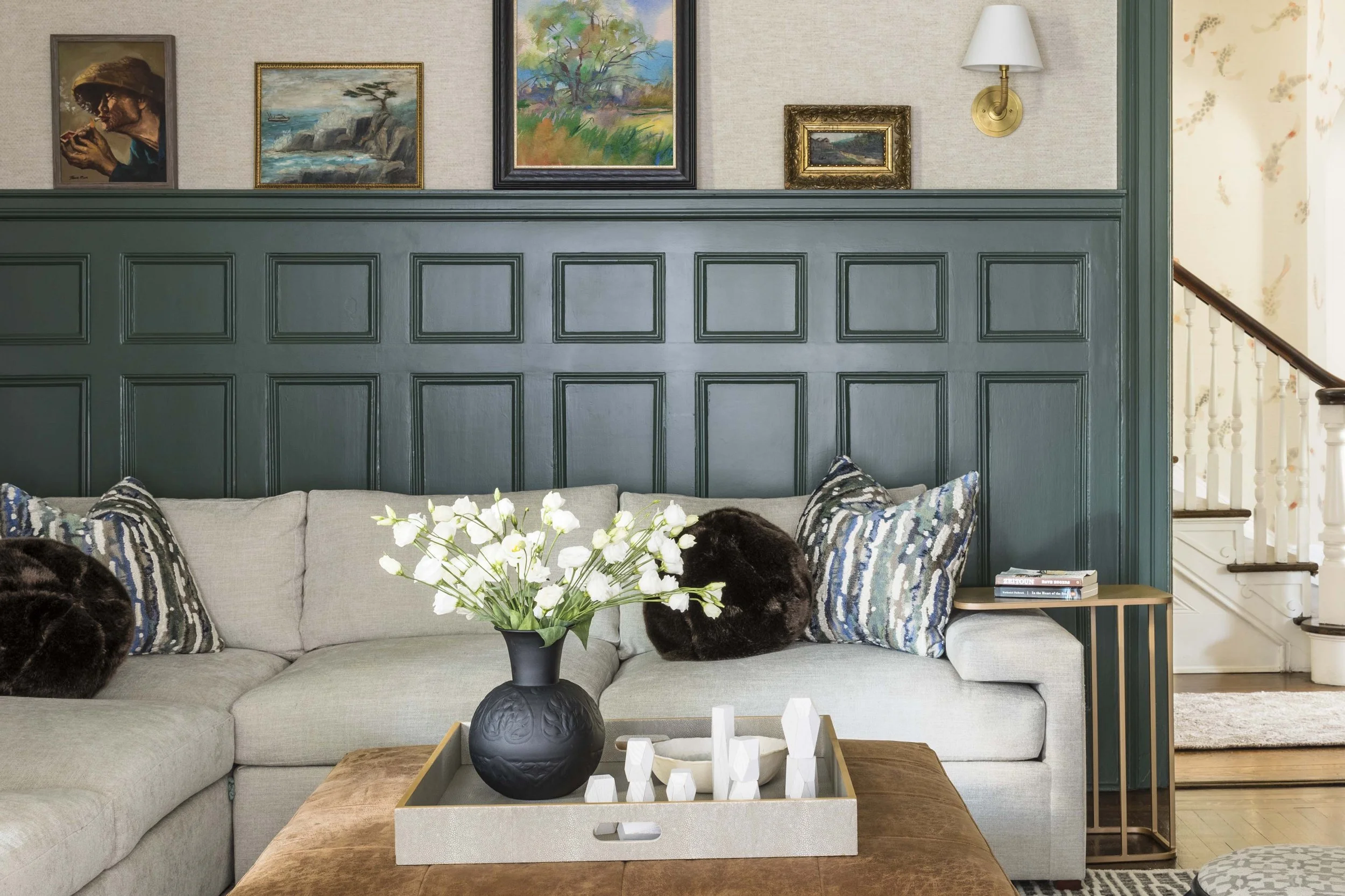 A living room with a beige sofa decorated with patterned pillows, a black vase with white flowers on a wooden coffee table, a gold side table with books, an artwork gallery on a green wainscoted wall, and a staircase in the background.