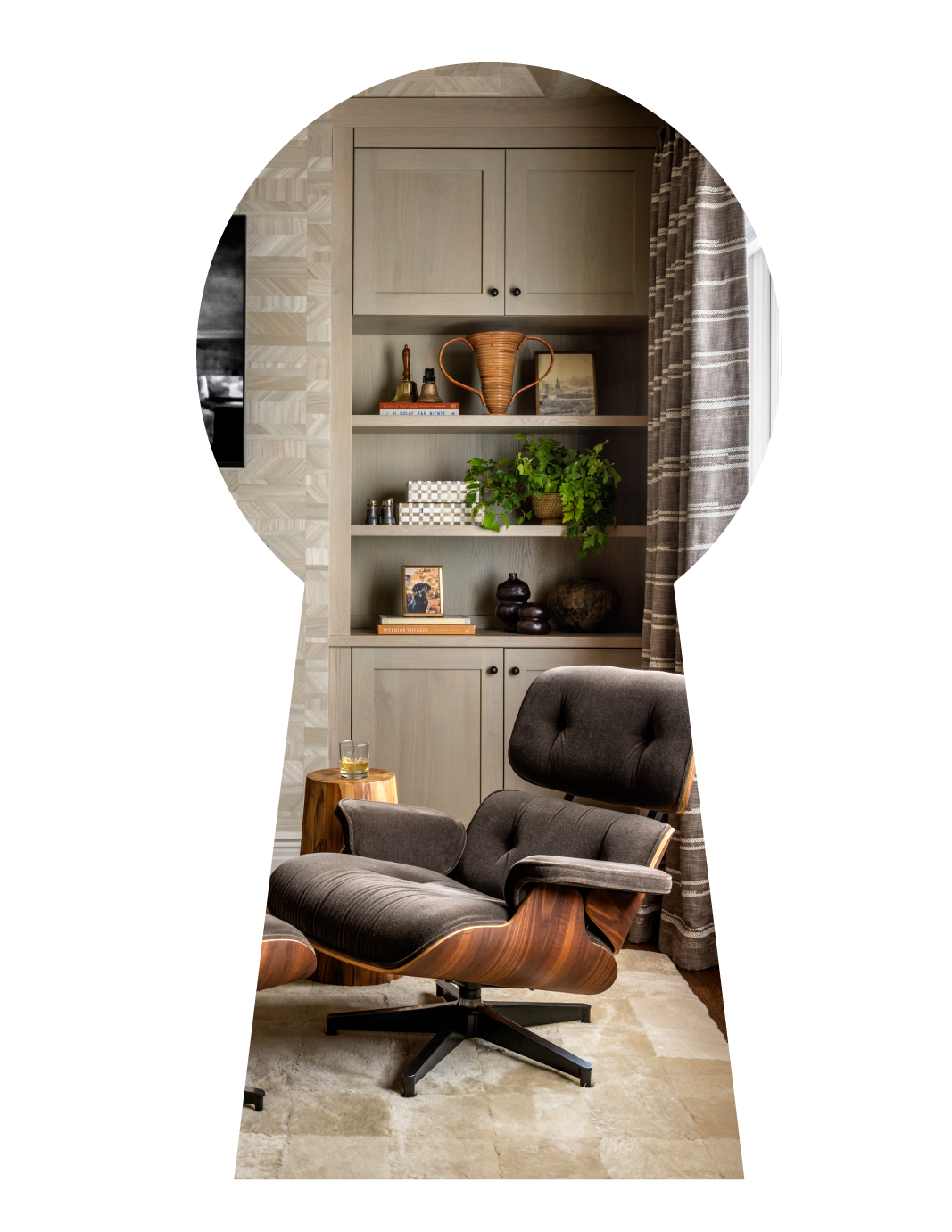 Interior of a cozy living space with a vintage lounge chair, a small wooden side table with a glass of beverage, a built-in bookshelf with decorative items and a plant, seen through an arched opening with patterned curtains and a large window.