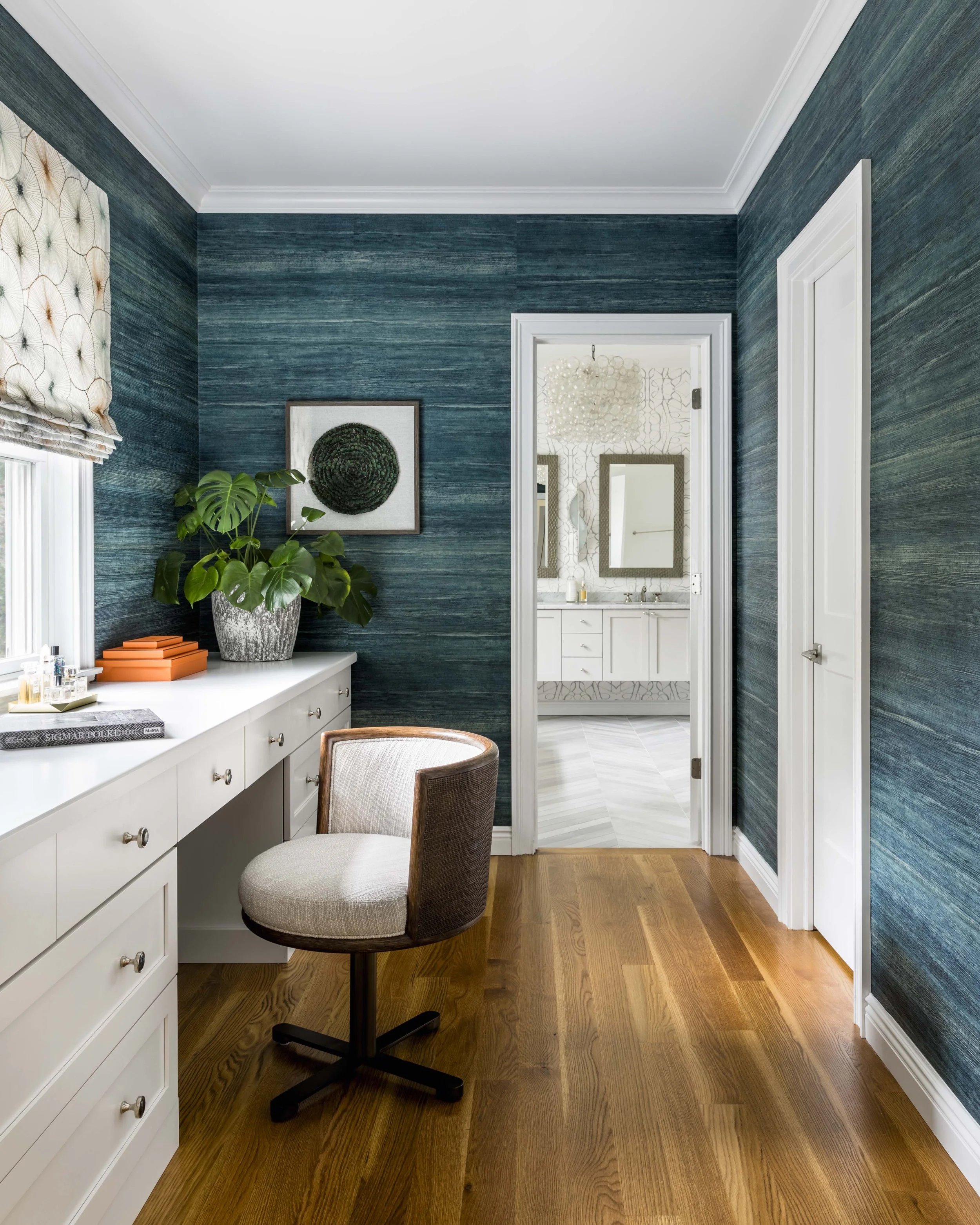 Home office with white desk, beige office chair, blue textured wallpaper, potted plant, framed wall art, window with Roman shade, connected to a bathroom with white vanity and mirrors.