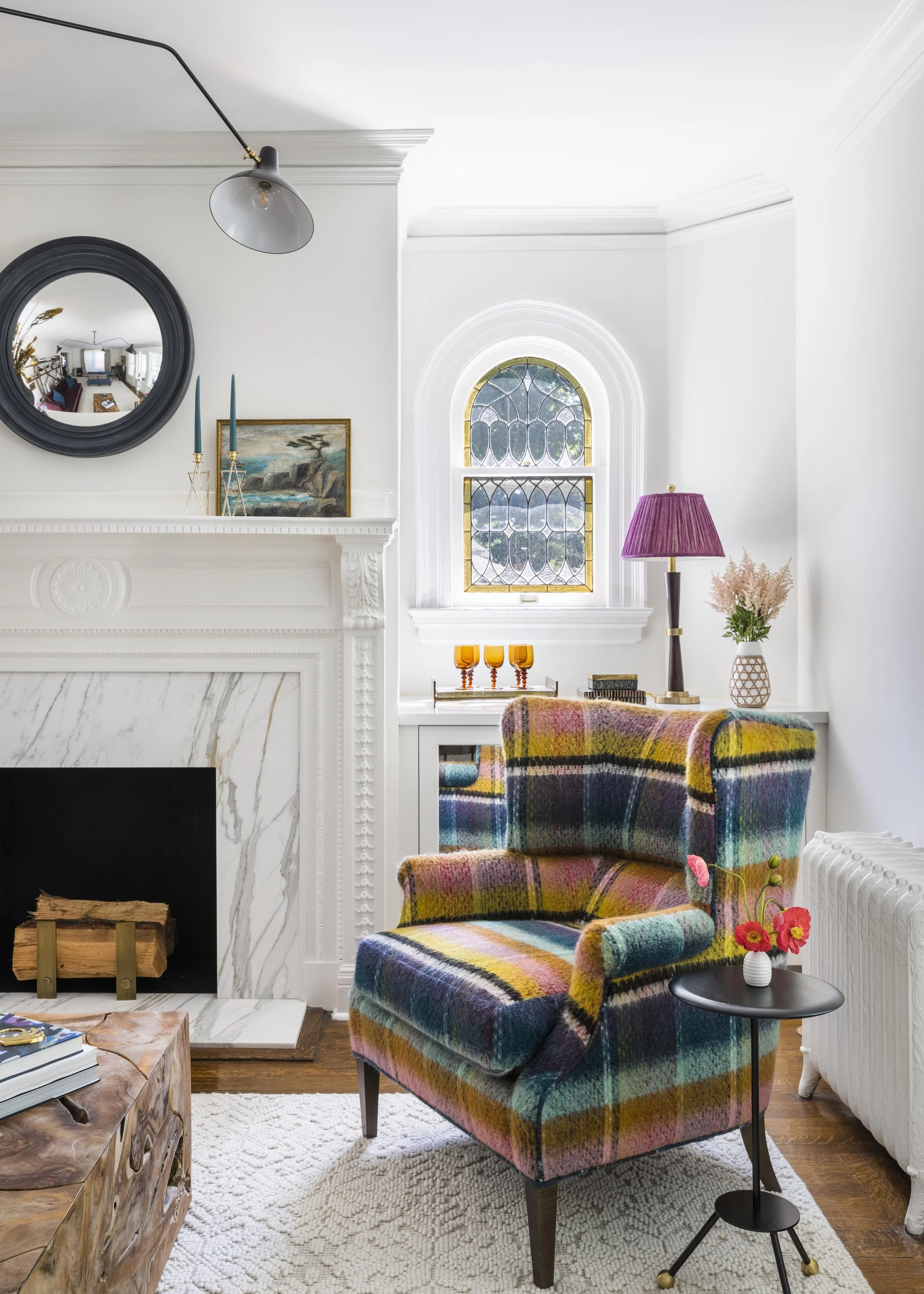 A colorful plaid armchair sits in front of a white fireplace with a black mantel. On the mantel are two teal candles, a small painting, and a round mirror. To the right of the armchair is a black side table with a white vase holding pink flowers. Behind the armchair, a window with stained glass allows natural light into the room. There is a pink floor lamp with a pleated shade and pink flowers on the table in the background.