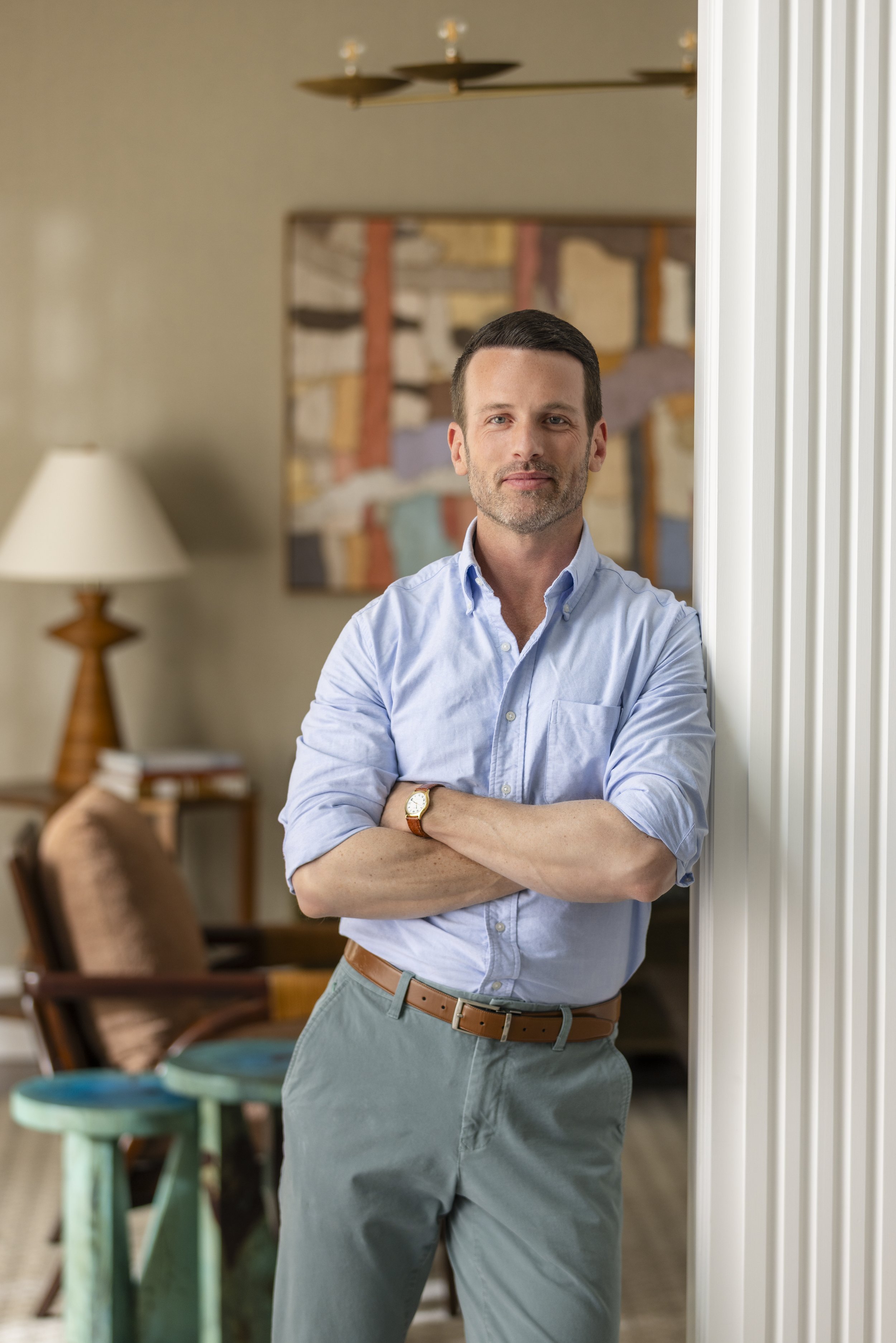 A man with short dark hair and a beard, wearing a light blue button-up shirt with sleeves rolled up, beige pants, and a brown belt, standing with arms crossed in a room with a couch, lamp, chair, and colorful abstract art on the wall.
