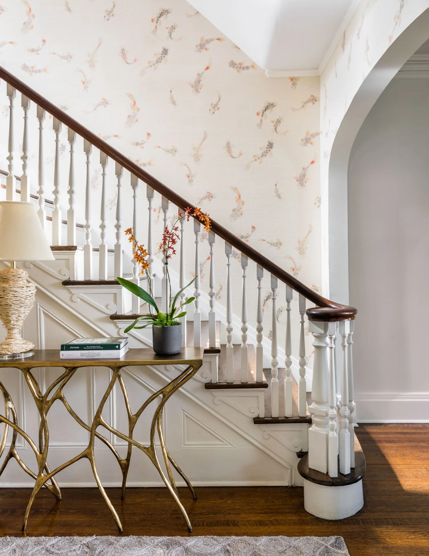 Interior view of a staircase with a wooden handrail and white balusters, a decorative side table with a gold-colored, organic design, a potted orchid plant, and pastel-colored wallpaper with a feather pattern.
