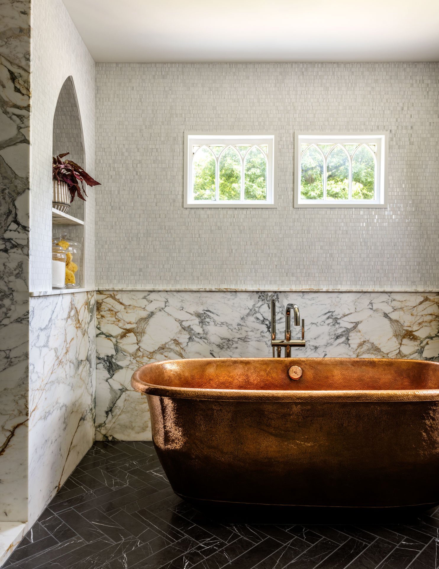 A bathroom features a copper bathtub, marble wall accents, two arched windows with a view of green trees, and a built-in shelf with decor items.