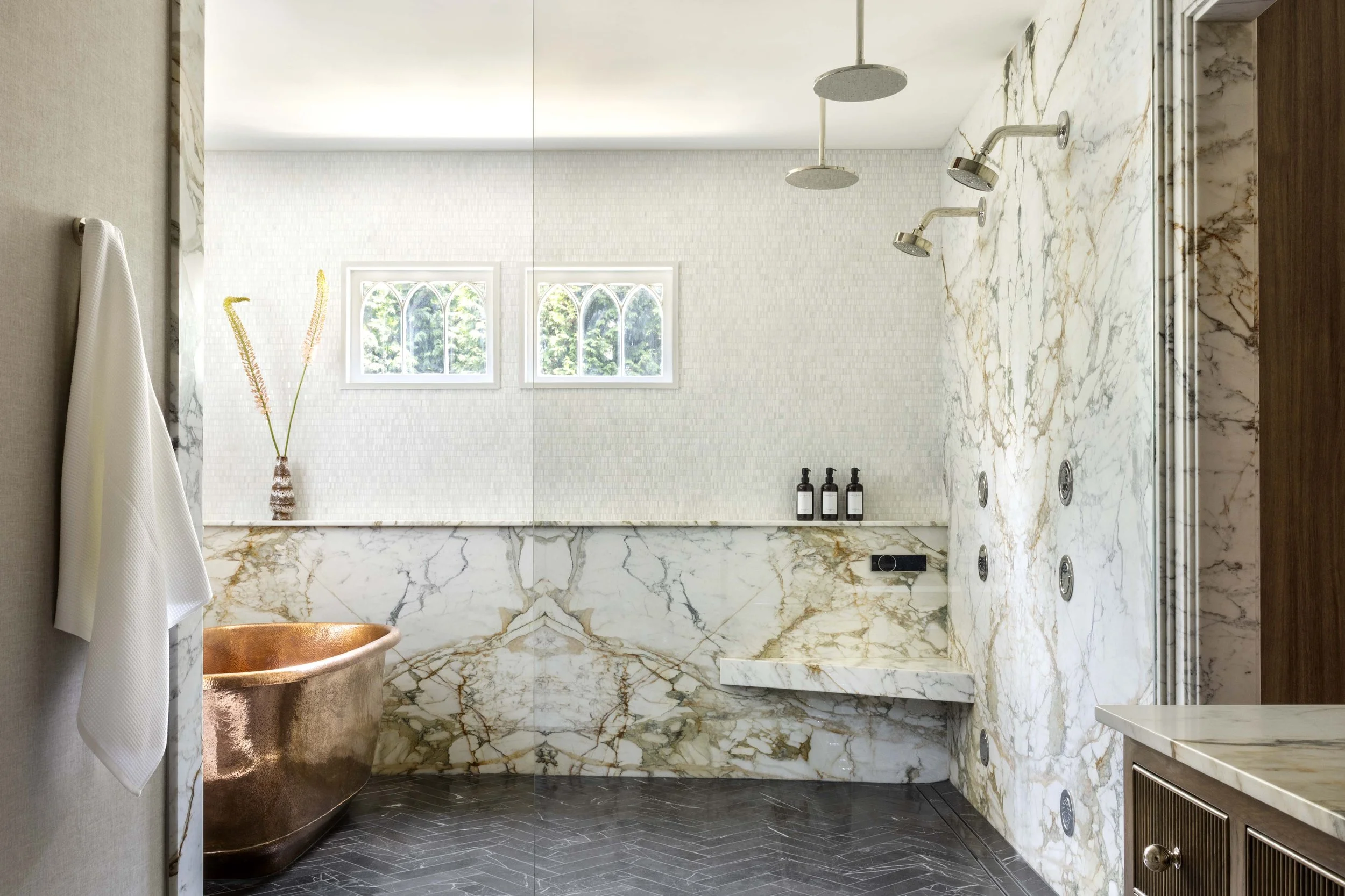 Bathroom with marble walls, copper bathtub, white textured wall, two arched windows, shower area with multiple showerheads, towel hanging on the wall, and a small cabinet with a marble countertop.