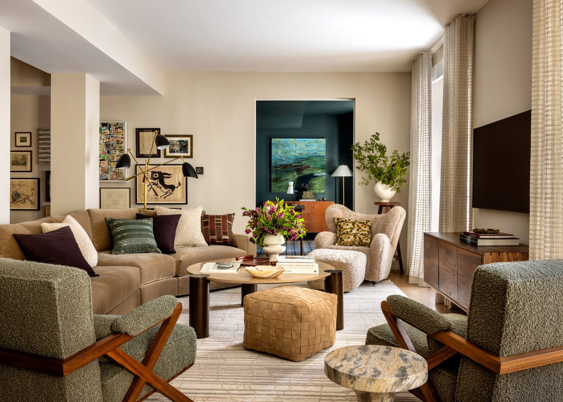 A cozy living room with a beige sectional sofa and two green armchairs, a round coffee table with a vase of flowers, and a patterned ottoman. Wall art and a large flat-screen TV are visible.