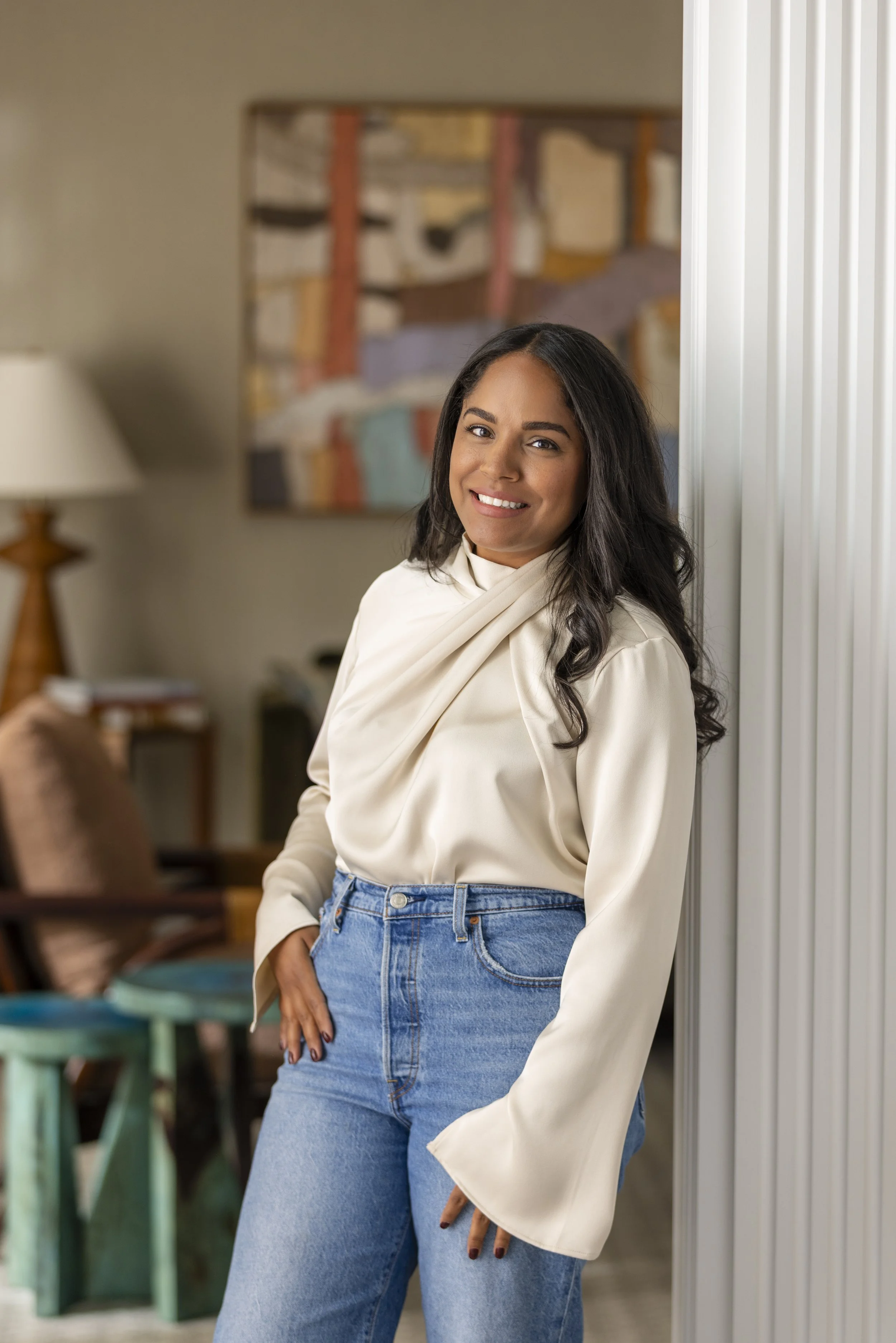 A woman with long dark hair, smiling and leaning against a wall in a cozy living room with colorful wall art and a side table with a lamp.
