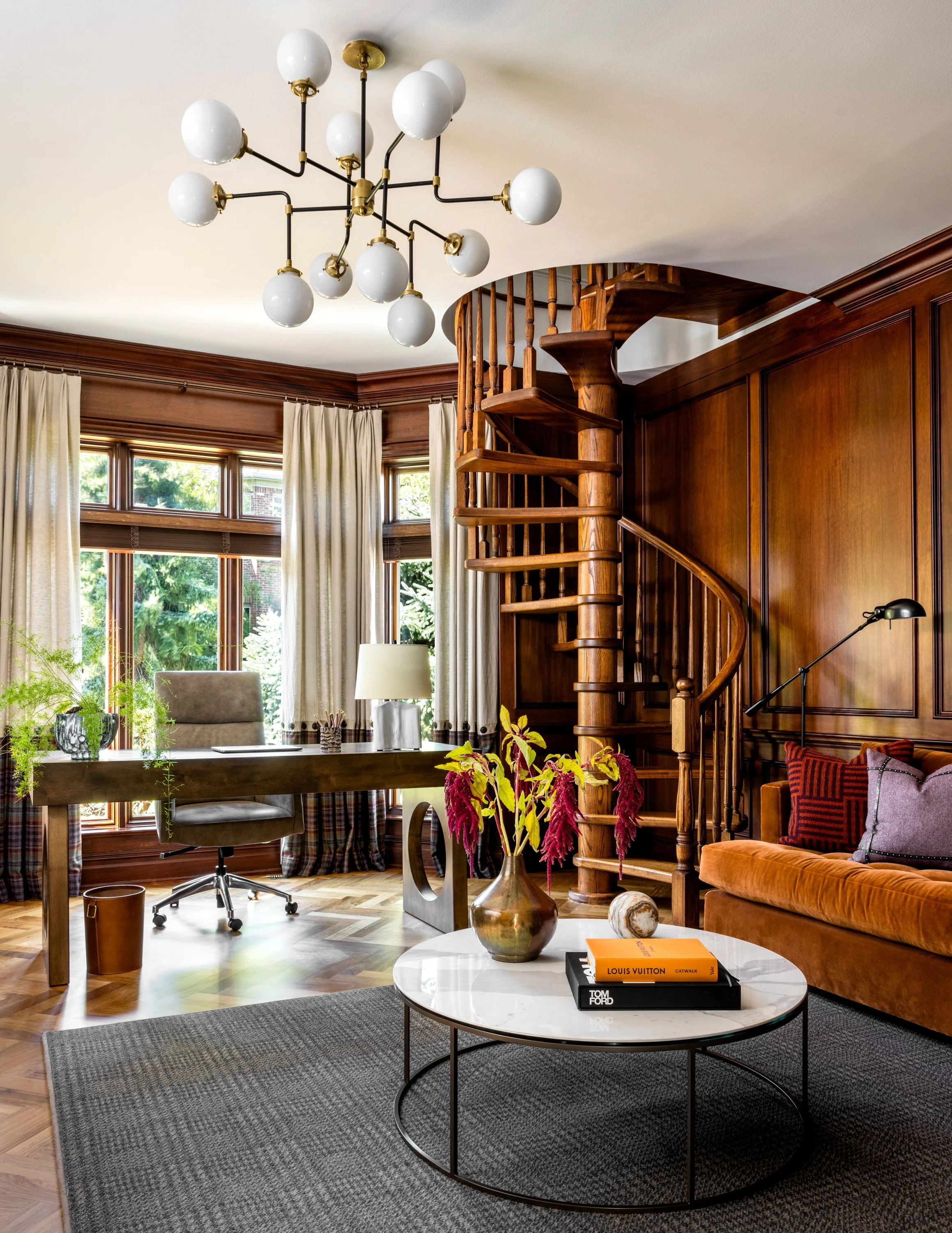 A cozy living room with a wooden spiral staircase, large windows with curtains, a desk with a chair, a lamp, a coffee table with a vase of pink flowers, books, pillows on an orange couch, and a modern chandelier hanging from the ceiling.