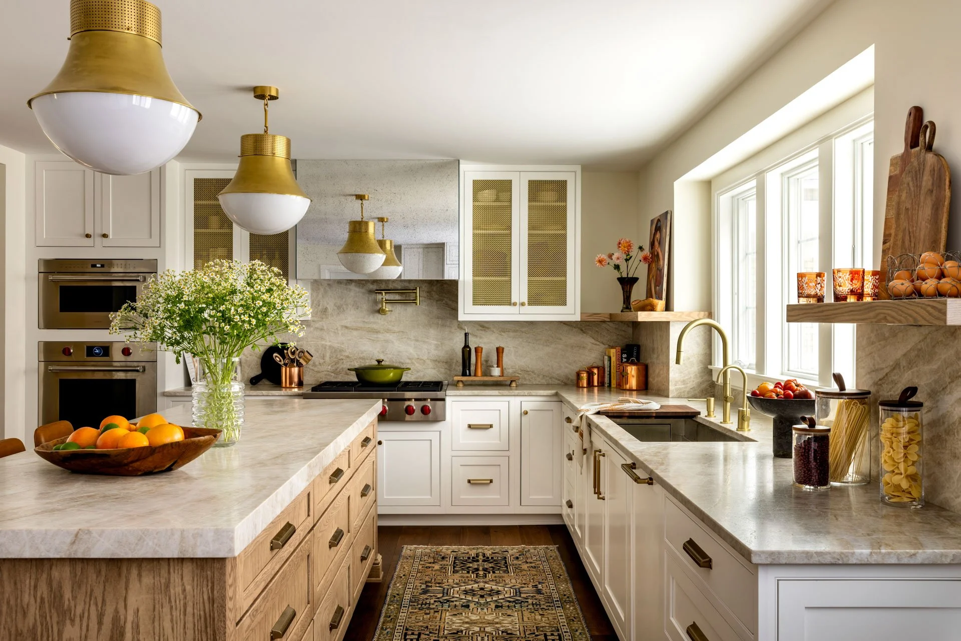 Bright kitchen with white cabinets, marble countertops, and gold fixtures. A large island with fruit bowl, flowers, and various kitchen utensils. Large window with shelf holding eggs and jars of pasta and beans.