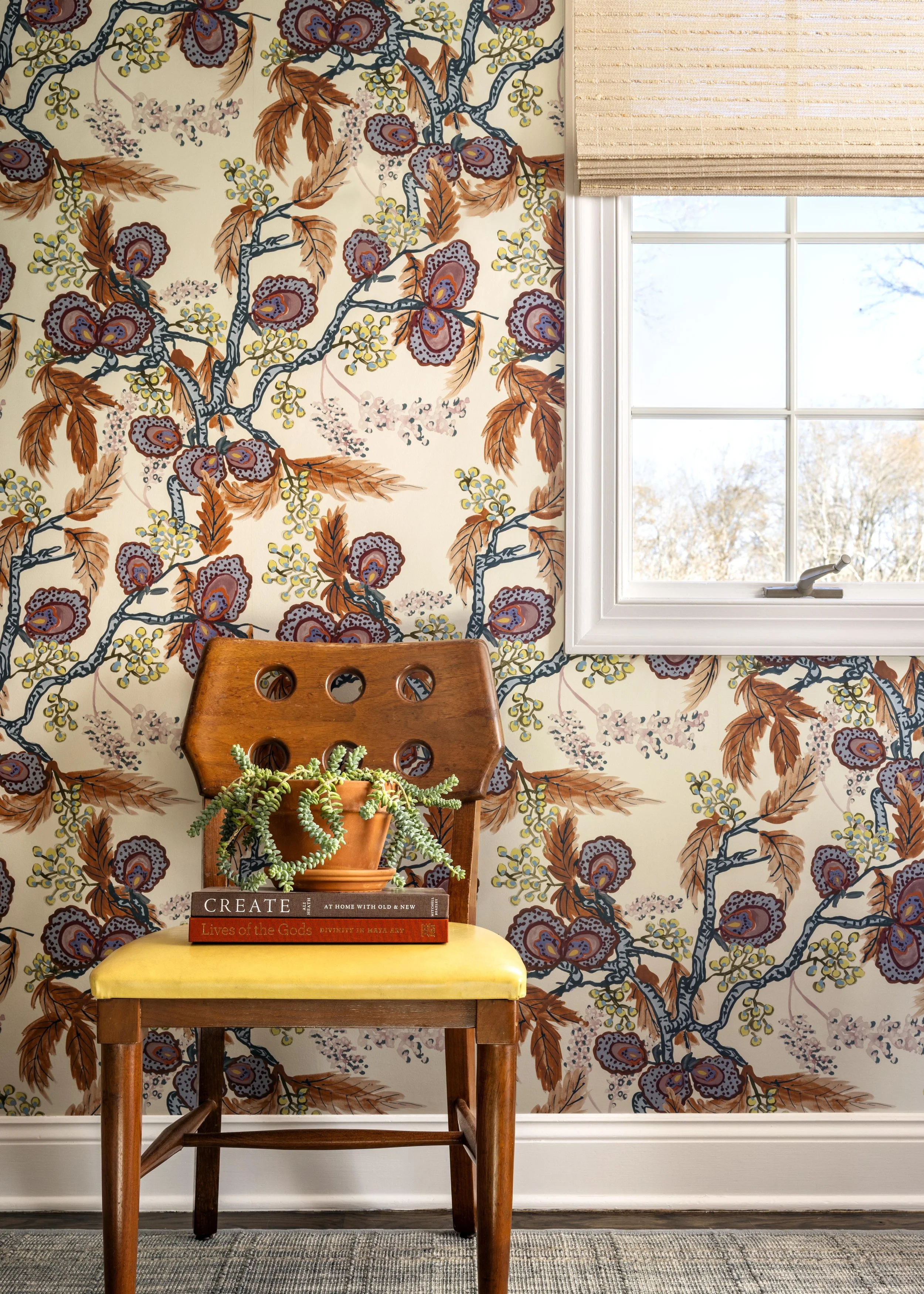 A wooden chair with a yellow cushion, a potted succulent, and two books on top, placed against wallpaper with a floral pattern of brown leaves, purple and blue flowers, and yellow accents, beside a window with beige blinds showing a tree outside.