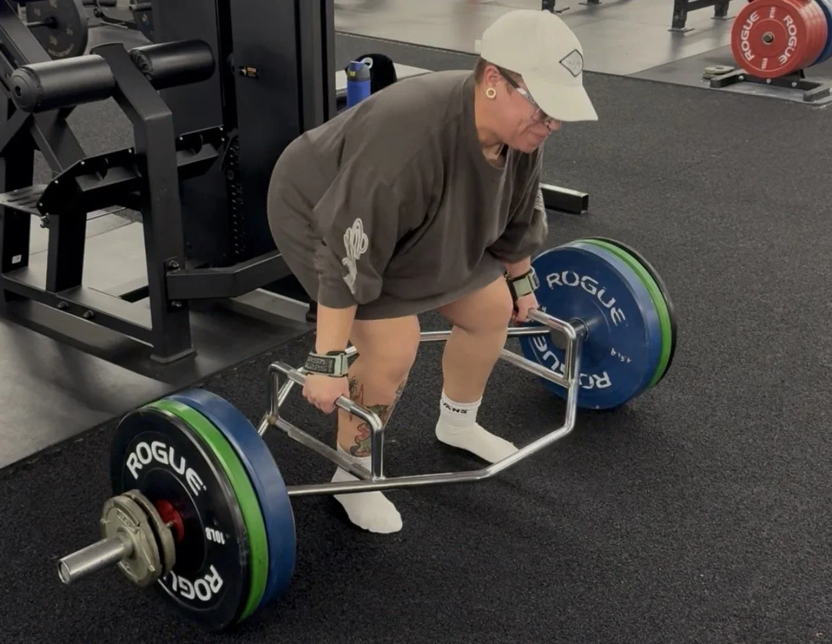 A woman in a gym lifting a barbell loaded with large weight plates, wearing a gray hoodie, white cap, and socks, with workout equipment and weight plates in the background.
