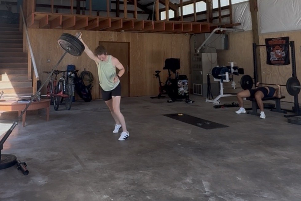 Two women working out in a gym with various exercise equipment and a wooden staircase in the background.