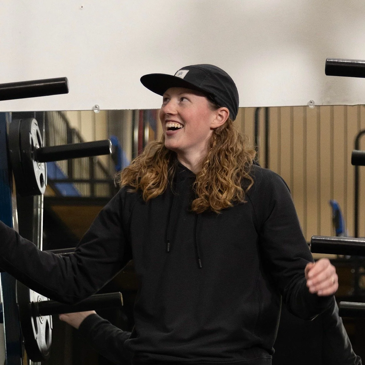 A woman with curly red hair wearing a black hoodie and black cap smiling at a gym.
