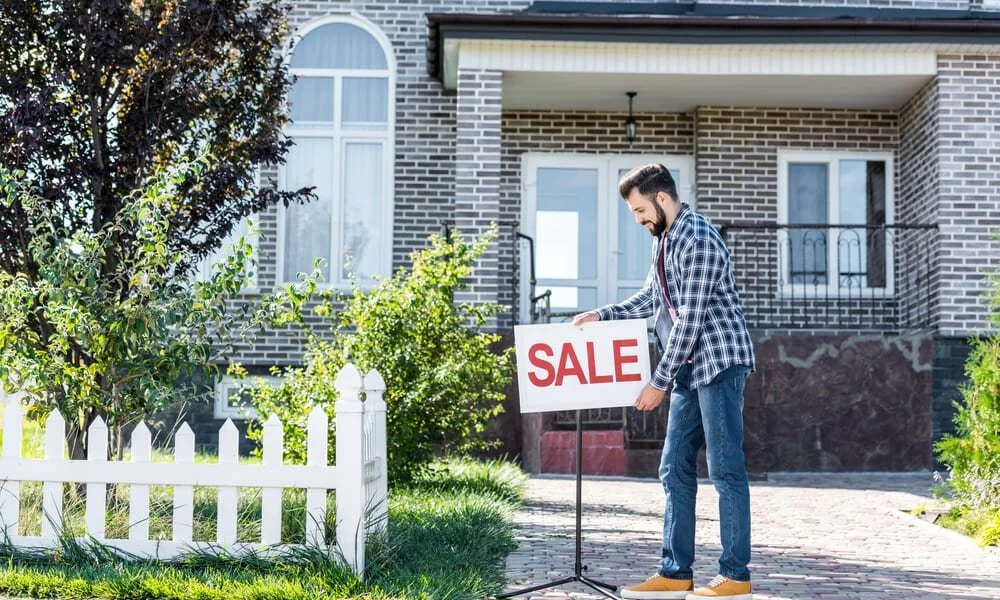 Man setting up a 'SALE' sign outside a house with a brick exterior and a small white picket fence.