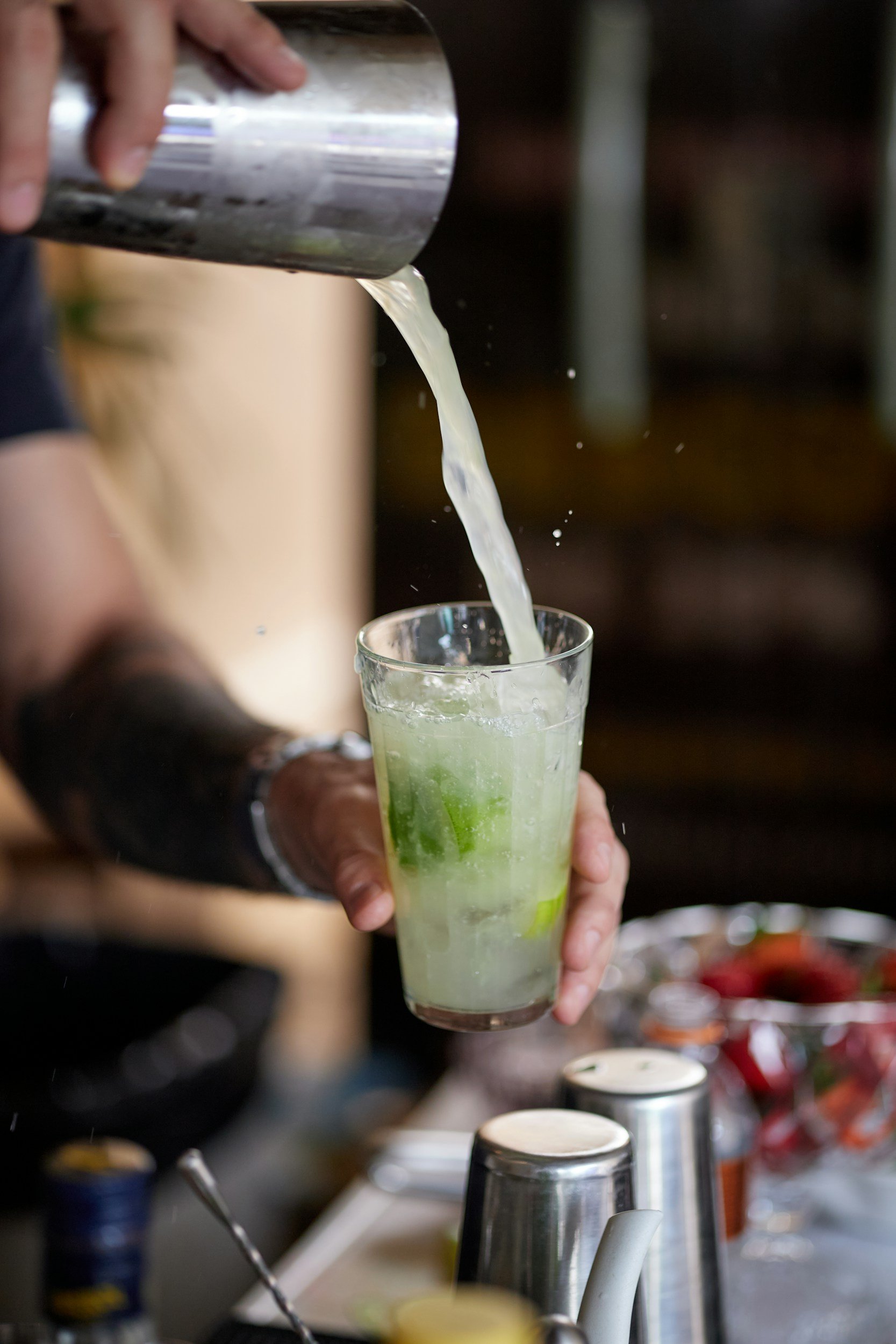 A row of pink cocktails garnished with edible flowers on a white tablecloth, with blurred people socializing in the background at an outdoor event.