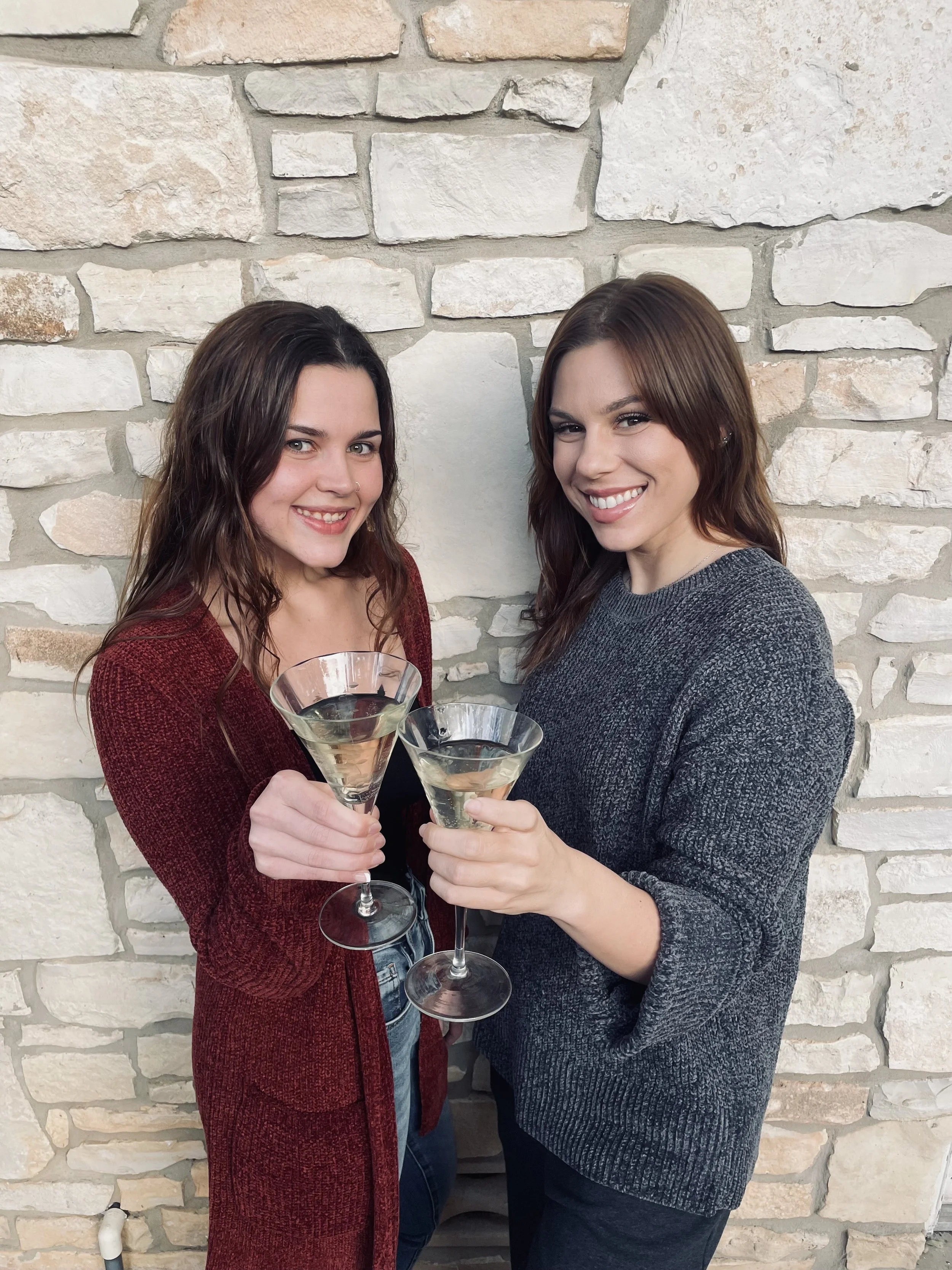 Two women smiling and holding glasses of wine in front of a stone wall.