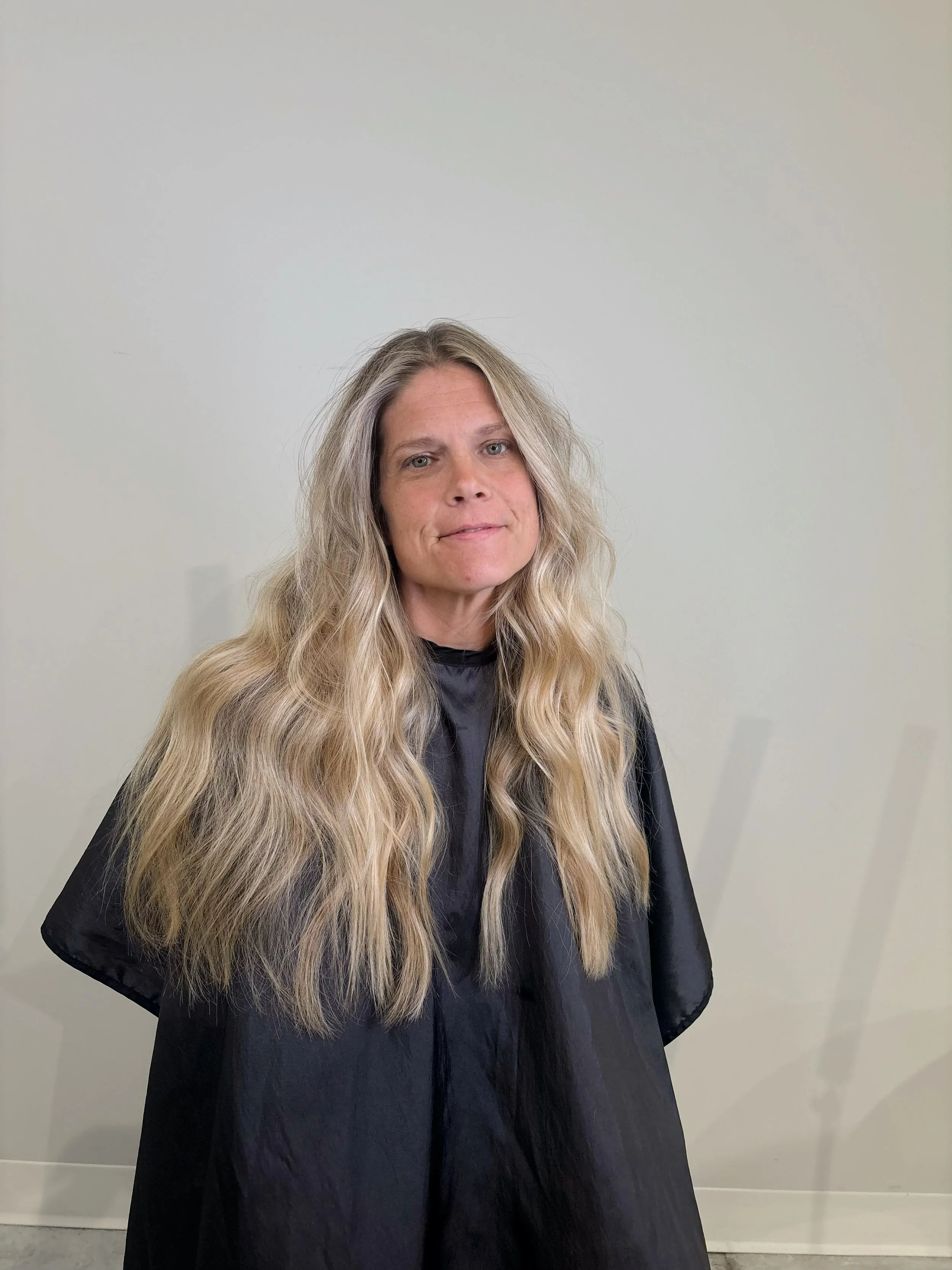 Woman with long wavy blonde hair wearing a black salon cape standing in front of a plain off-white wall.