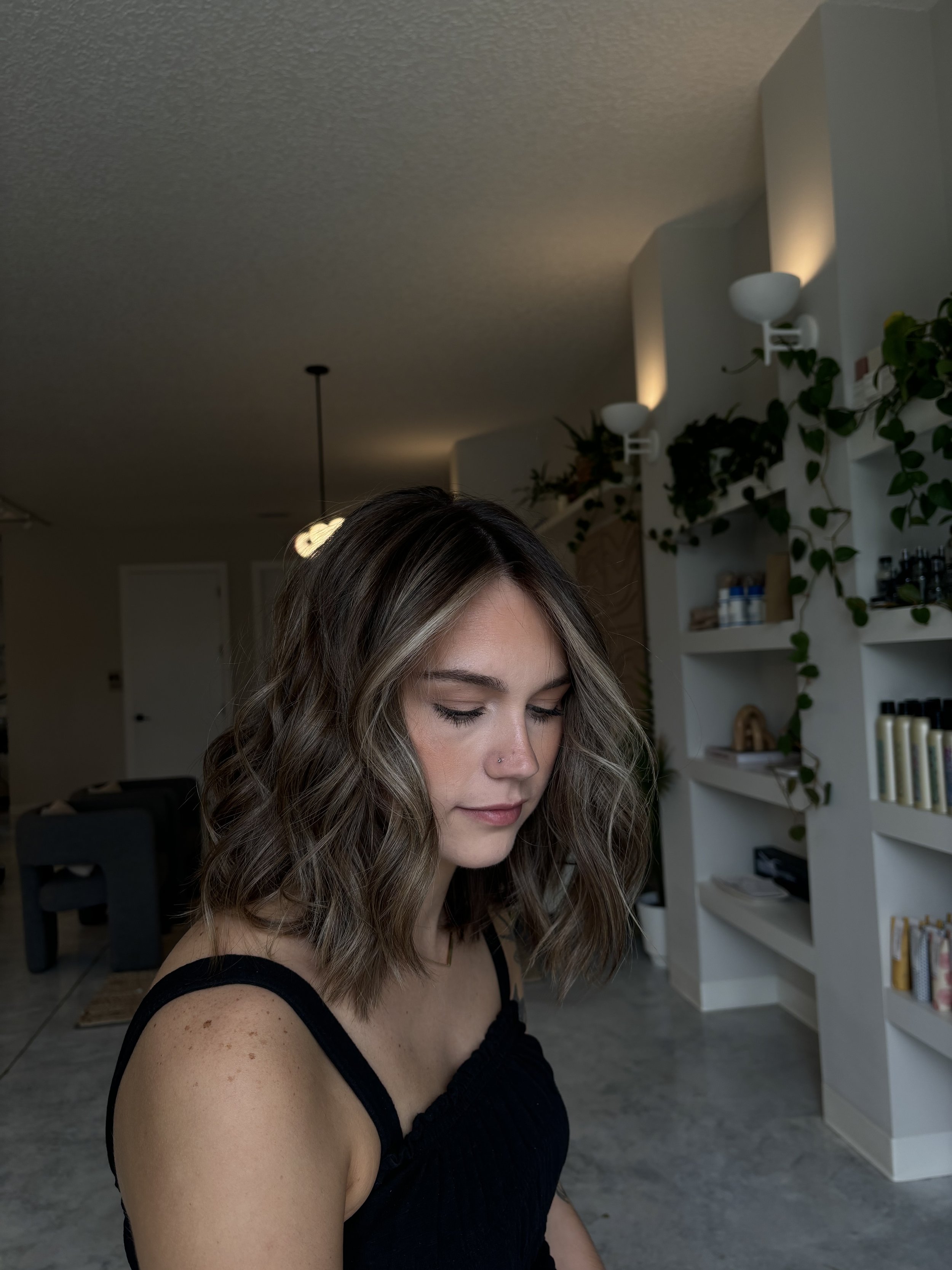 A young woman with shoulder-length wavy brown hair and a nose piercing, wearing a black sleeveless top, looking downward in a modern indoor setting with white shelves and green plants in the background.