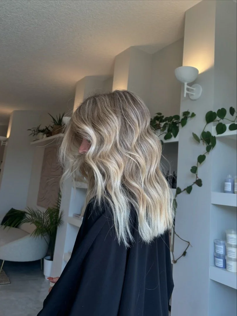 Side view of a woman with wavy blonde hair standing indoors with plants and furniture in the background.