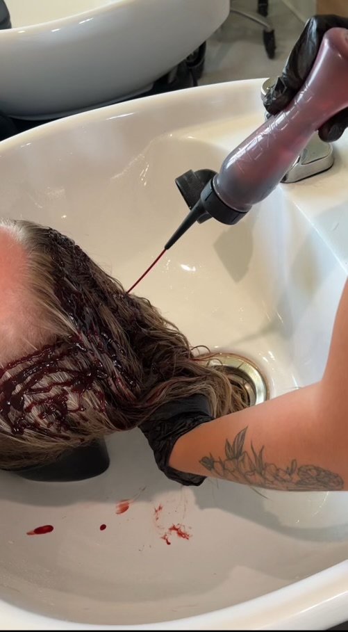 Hairdresser is rinsing a client's long, dark hair with a ceramic color in a salon sink.