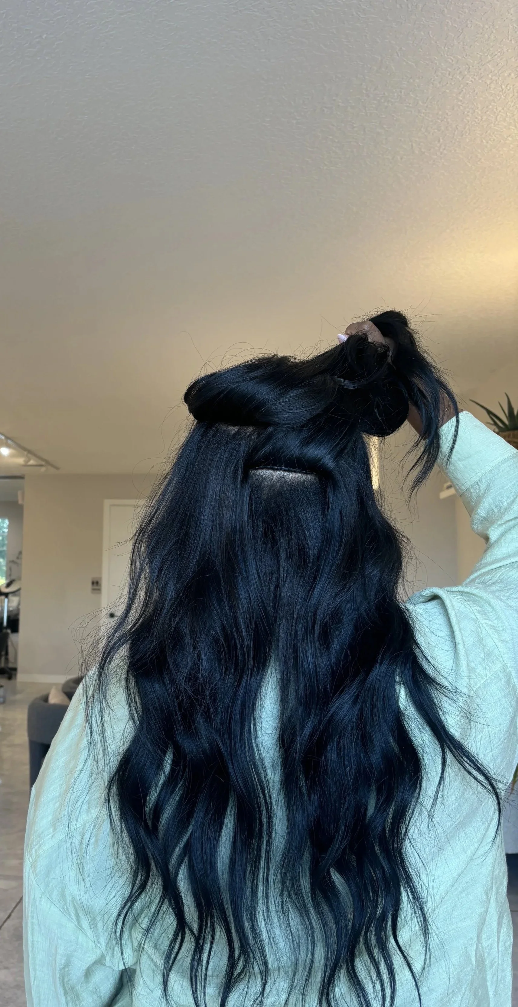 A person with long, wavy, black hair is seen from the back, holding up a portion of their hair with one hand, in a well-lit room.
