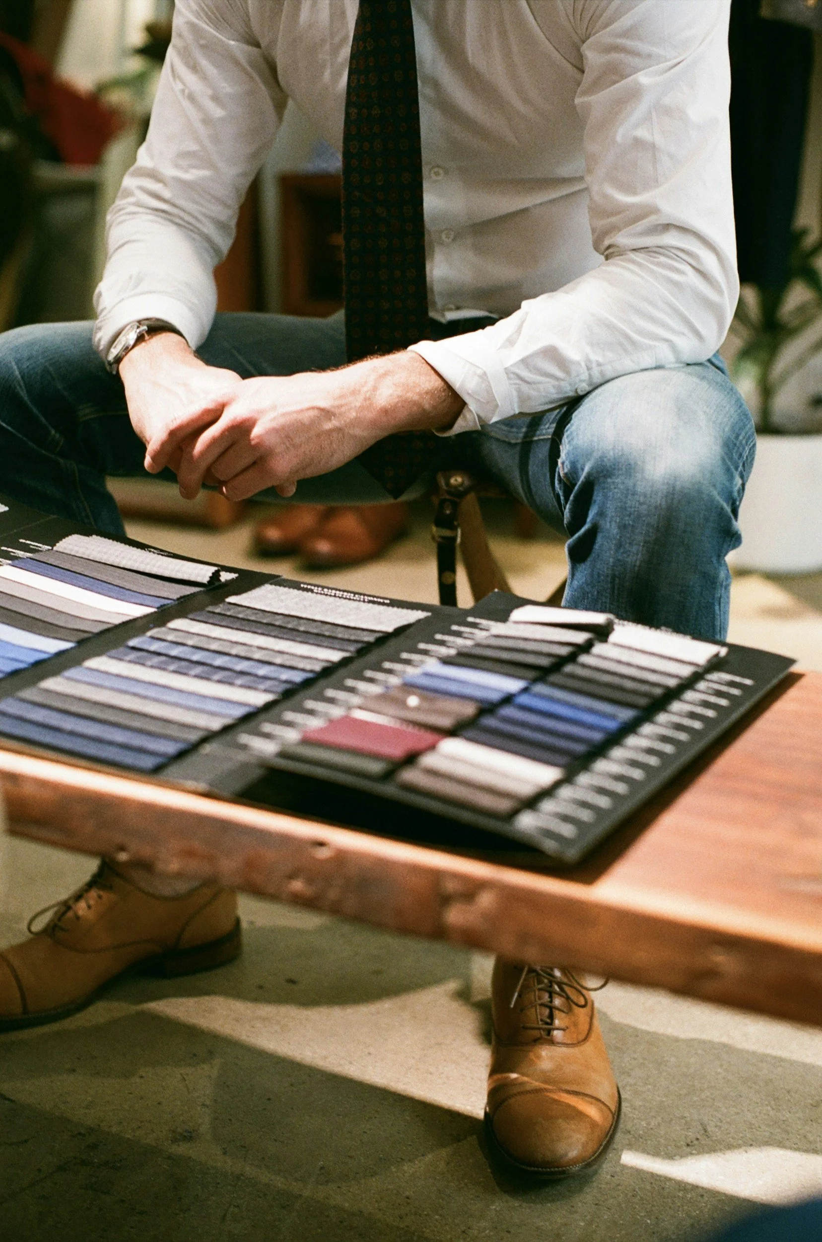 Person sitting at a table with fabric swatches, wearing jeans, a white shirt, and brown shoes.