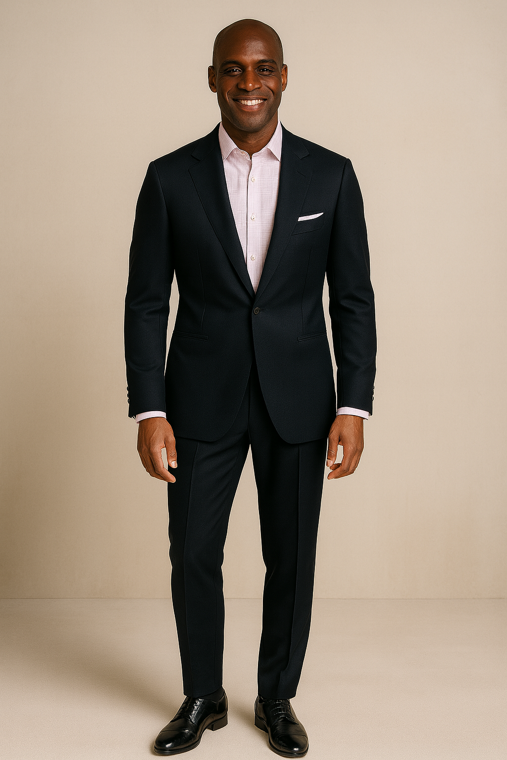 A man in a black suit with a pink shirt and black dress shoes, smiling while standing against a beige background.