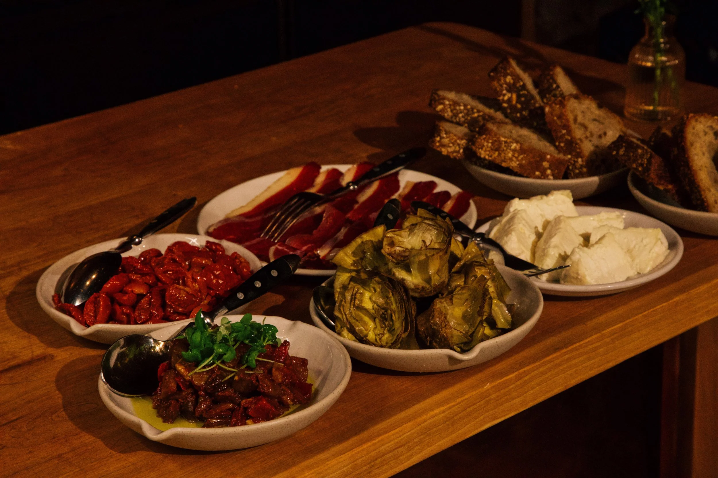 An assortment of Mediterranean appetizers on a wooden table, including roasted artichokes, sun-dried tomatoes, roasted peppers, slices of bread, slices of cured meat, slices of cheese, and a bowl of mixed greens, with small serving spoons in each dis