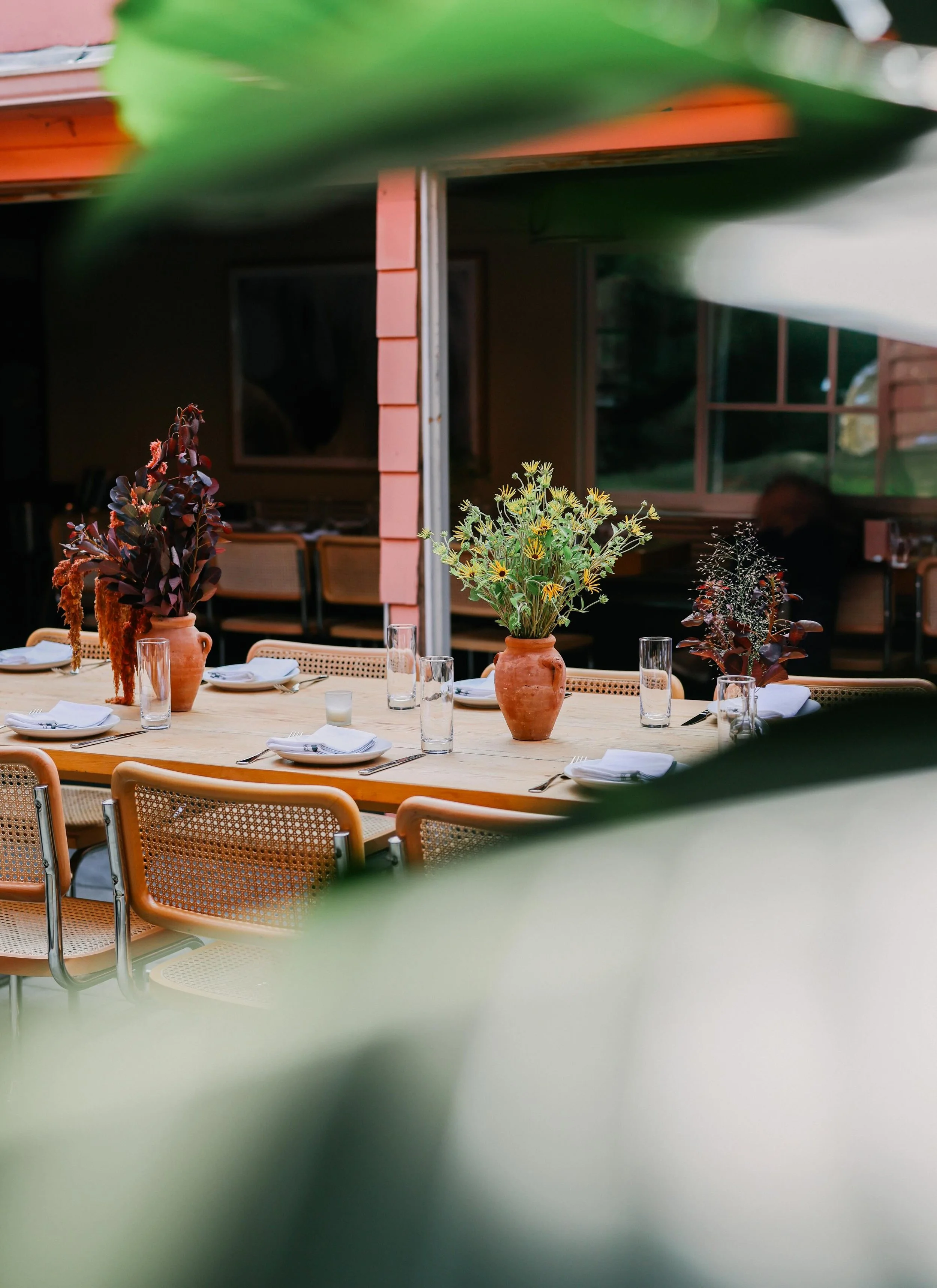 Decorated dining table with floral centerpieces, set with white plates, napkins, glasses, and silverware, viewed through blurred green leaves.