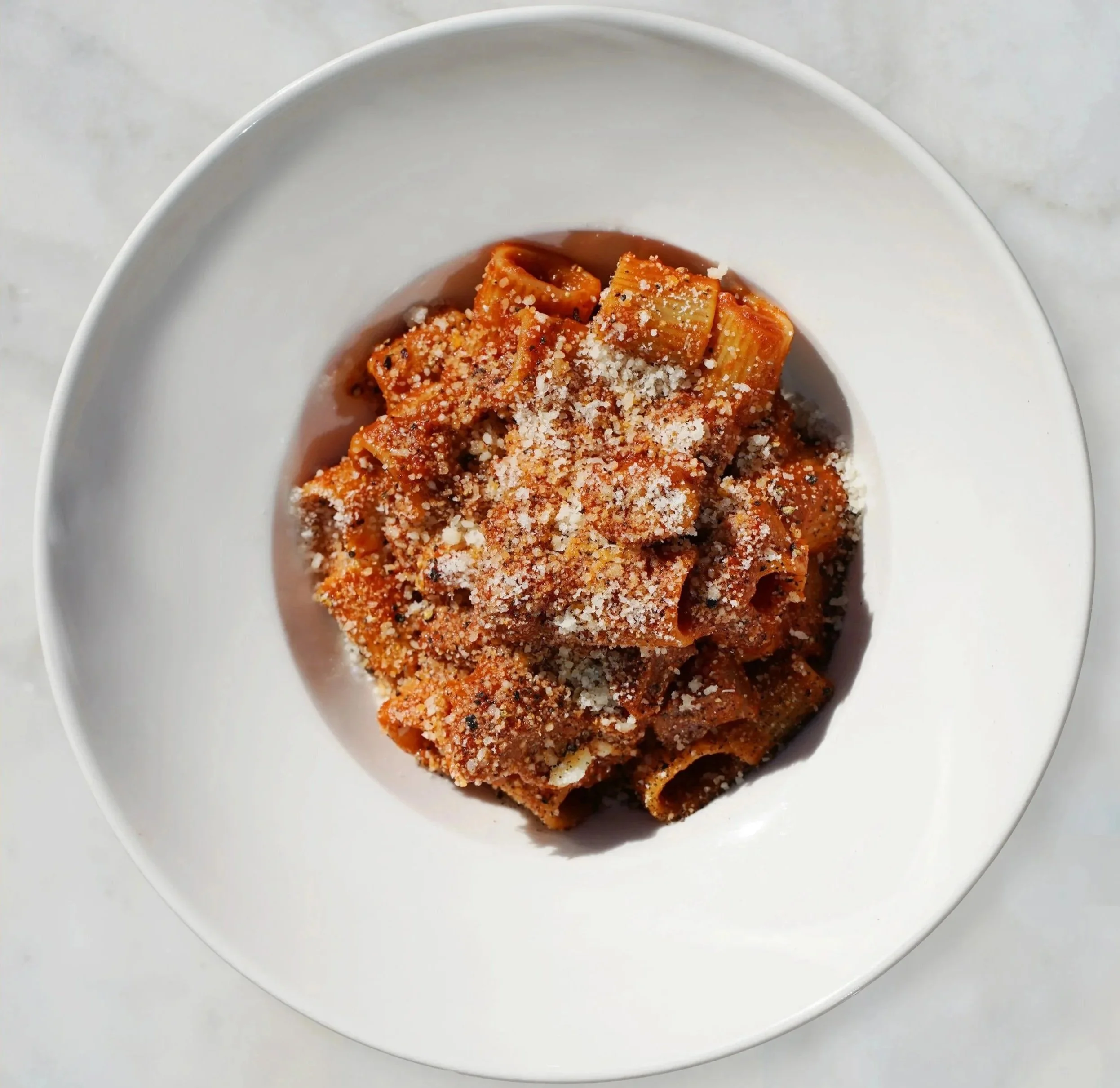 A white plate of cooked rigatoni pasta topped with tomato sauce and grated cheese at Roscioli NYC in the heart of SoHo.