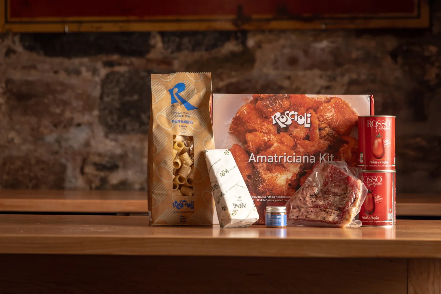 Italian cooking ingredients on a wooden surface, including a package of mezze maniche pasta, a box of roscali amatriciana kit, two cans of tomato purée, a sealed piece of pork, a small spice jar, and a wrapped block of cheese, with a rustic brick wall background.