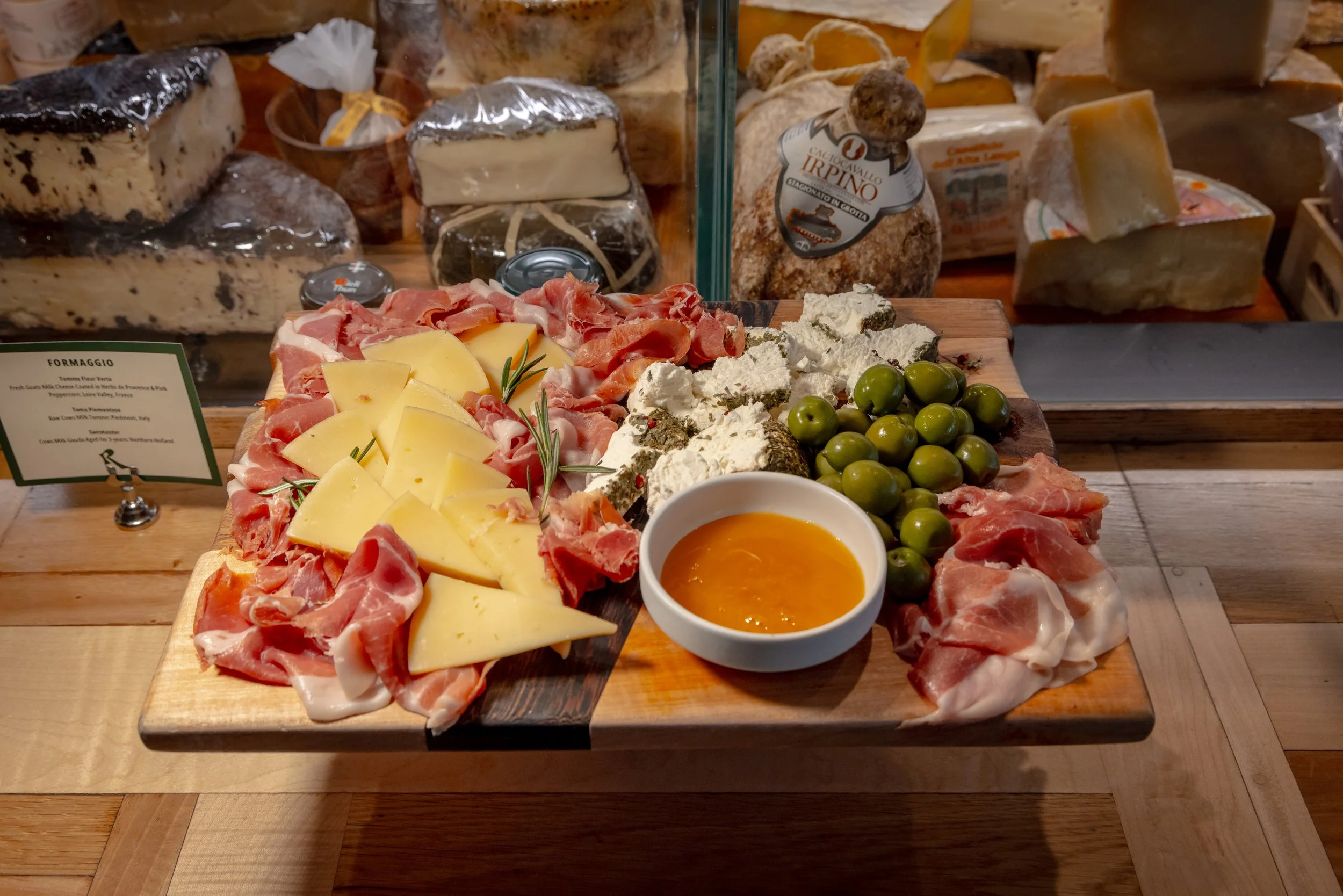 A wooden board with assorted cheeses, green olives, sliced cured meats, and a small bowl of honey or mustard, presented as a charcuterie platter.