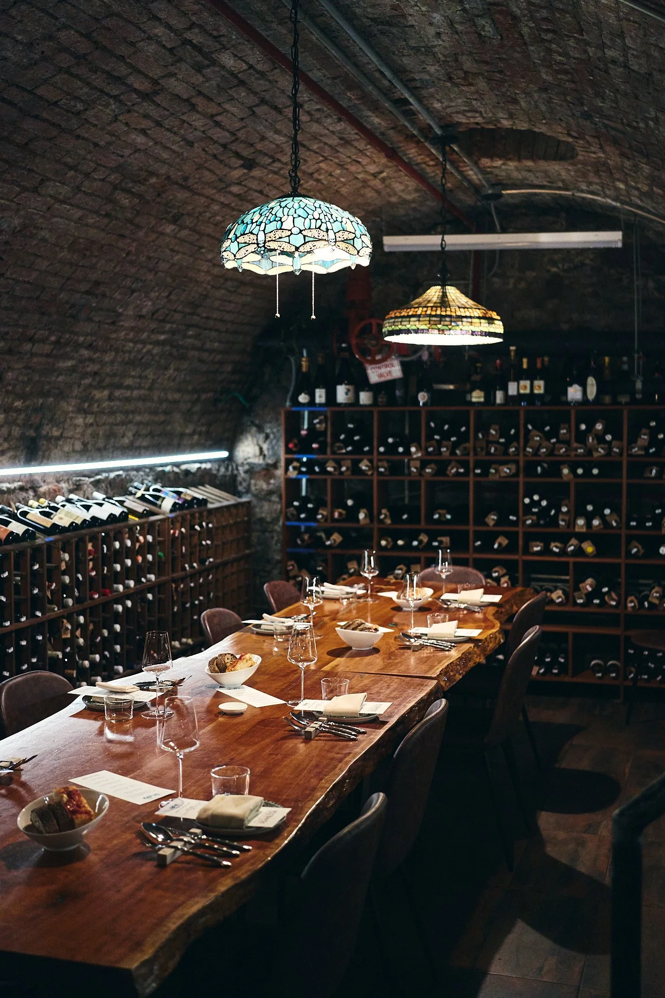 A cozy wine cellar inside Roscioli with a wooden dining table set for a meal, surrounded by wine bottles and shelves filled with wine bottles on a brick arched ceiling, illuminated by stained glass pendant lights.
