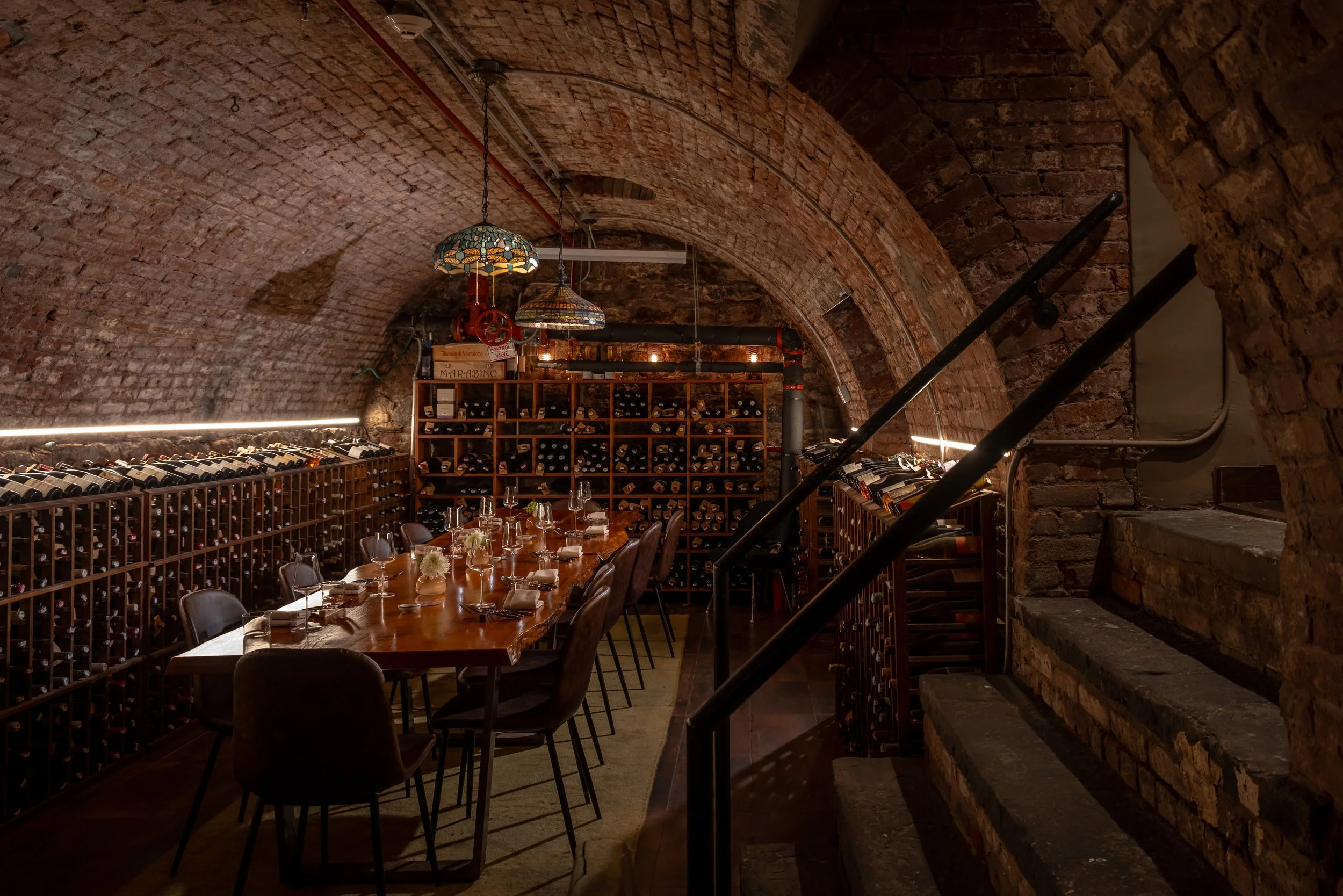A cozy wine cellar with brick arched ceiling, wine bottles stored on racks along the walls, a long wooden dining table set with glasses and flowers, and vintage hanging lamps.