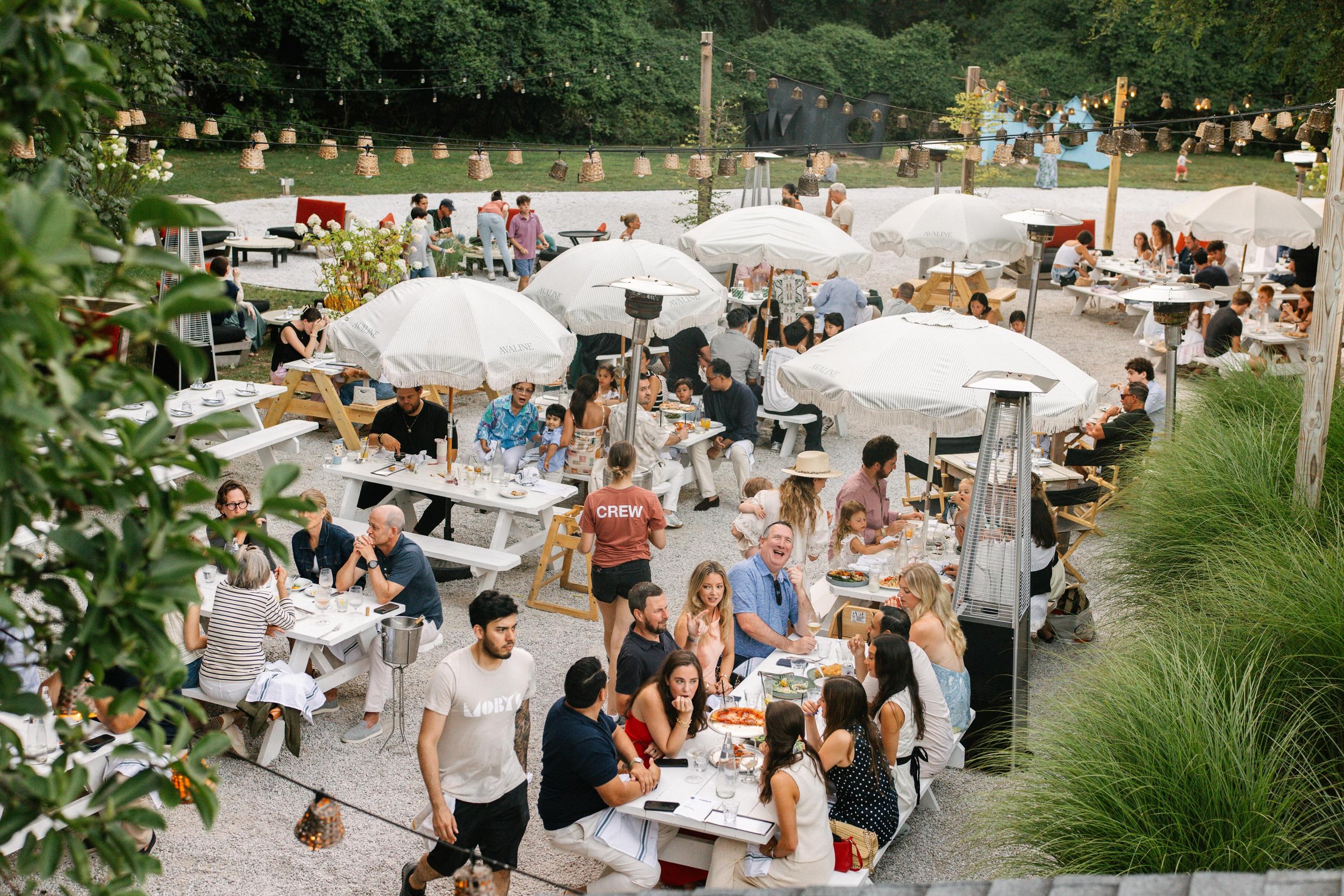 Outdoor catering event with people dining at tables and under umbrellas, string lights hanging overhead, and a gravel ground surrounded by greenery.