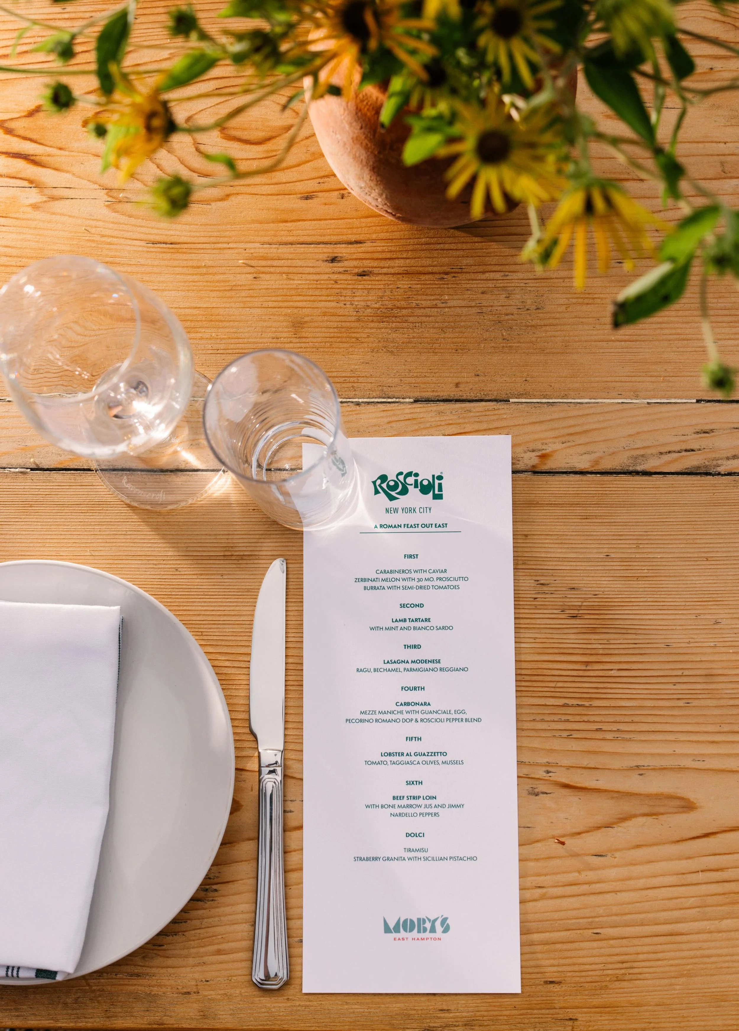 A restaurant table setting with a wooden table, a white plate with a folded white napkin, a knife, two empty glasses, a menu, and a potted plant with yellow flowers.