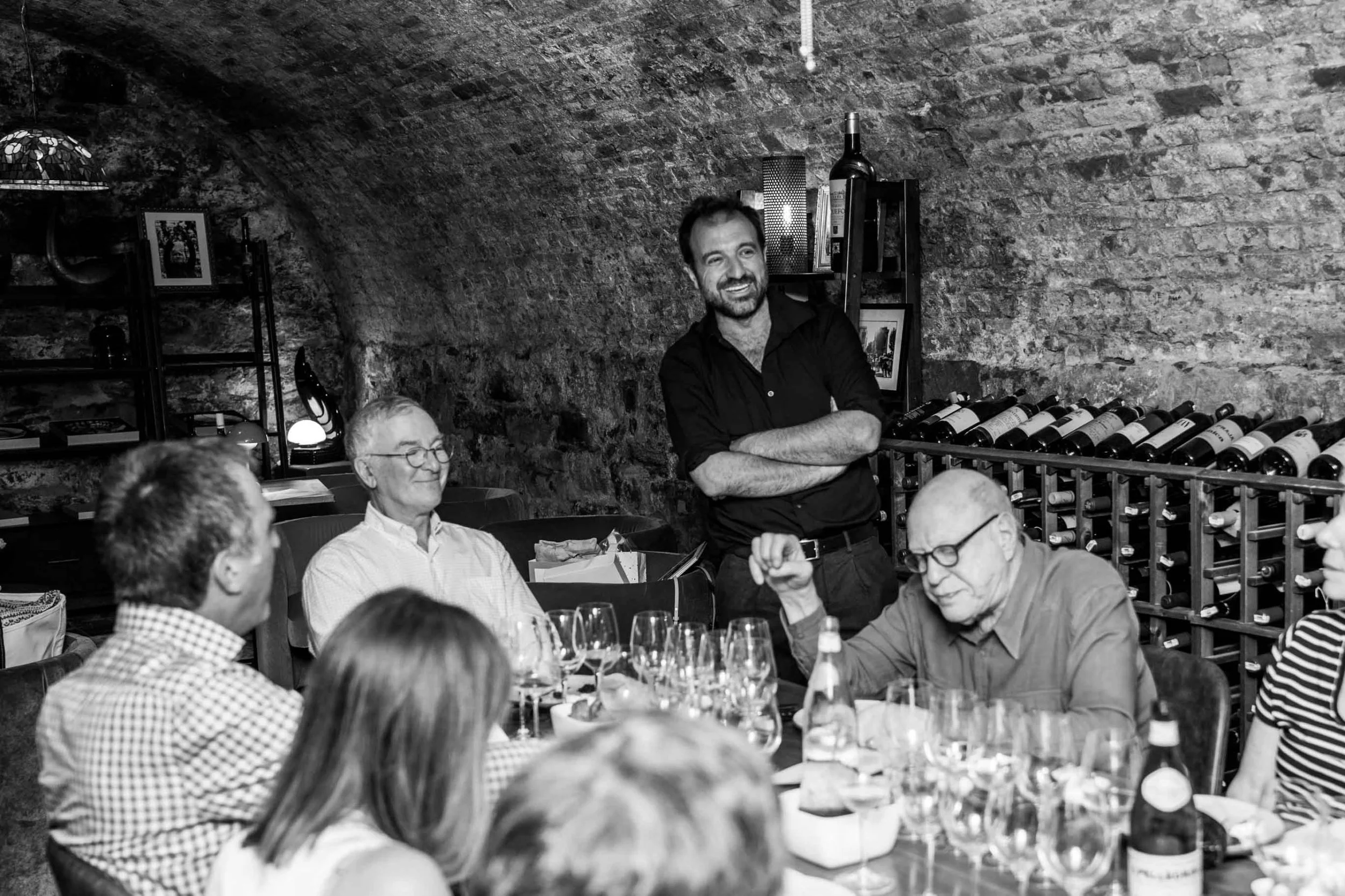 A group of people sitting around a table in Roscioli NYC's wine cellar, with a man standing and smiling near wine bottles on a rack against a brick wall.