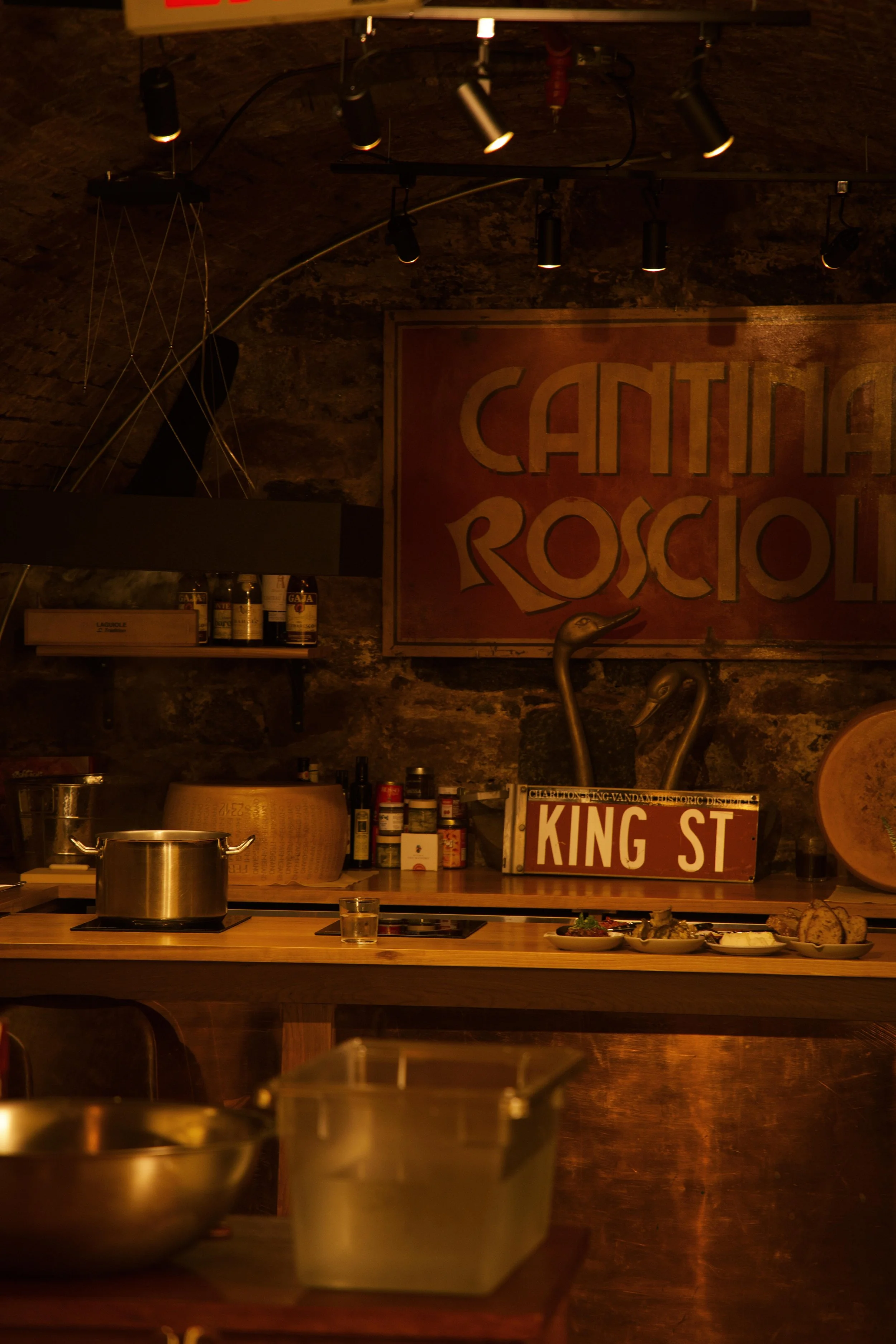Interior of Roscioli's rustic restaurant kitchen with a stone wall, warm lighting, shelves with bottles, a stainless steel pot on the counter, where cheese and wine classes are held.