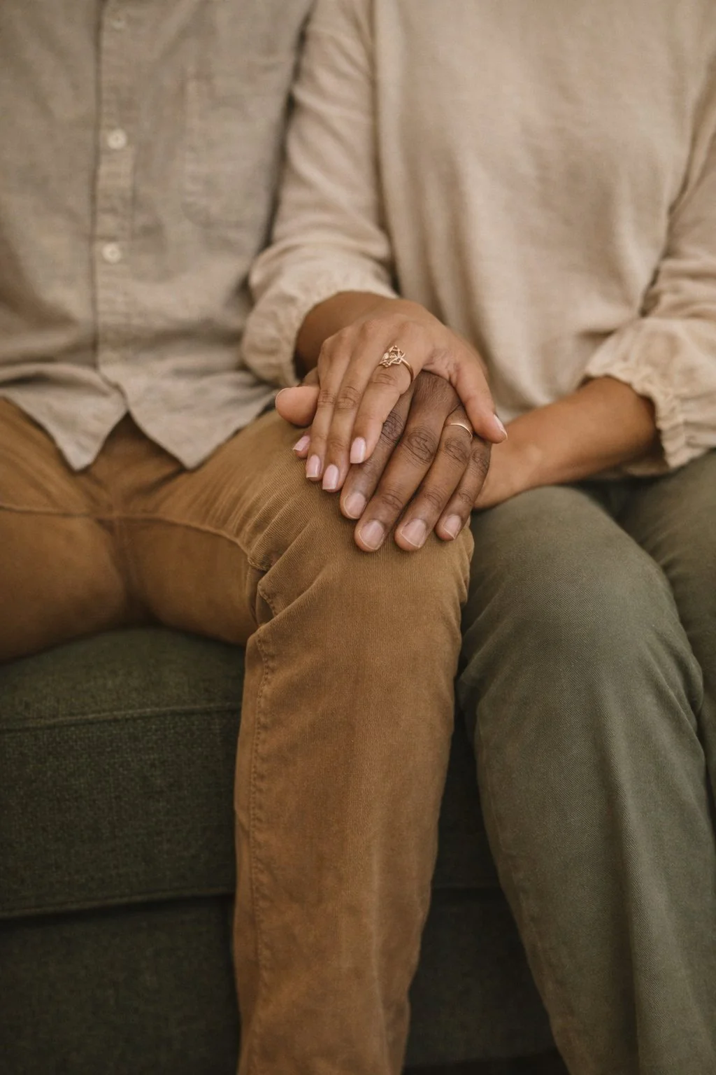 A couple sitting on a sofa, holding hands on their laps, with the woman's hand on top showing a gold ring, and the man wearing a ring on his finger.