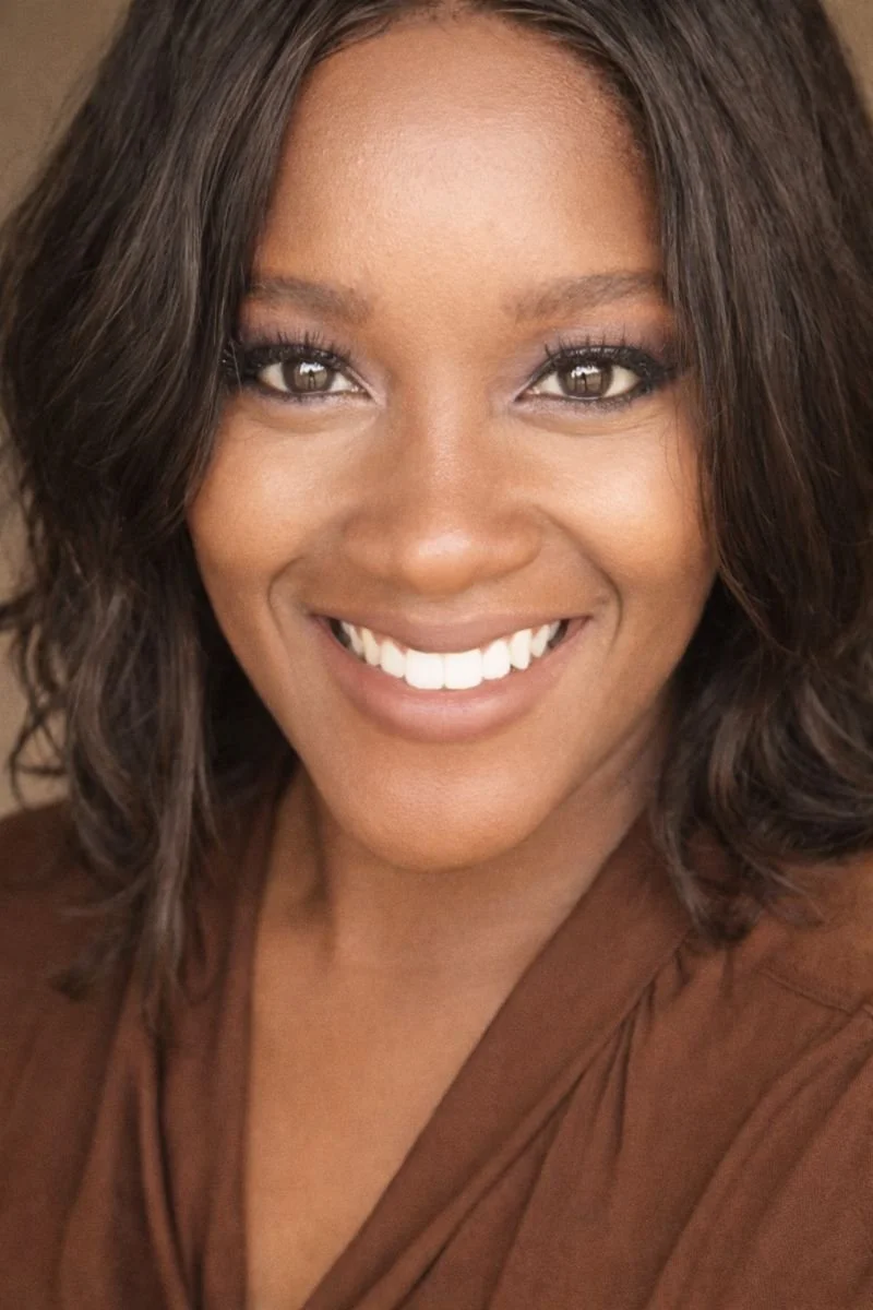 Close-up of a smiling woman with dark, wavy hair and brown eyes, wearing a brown top.