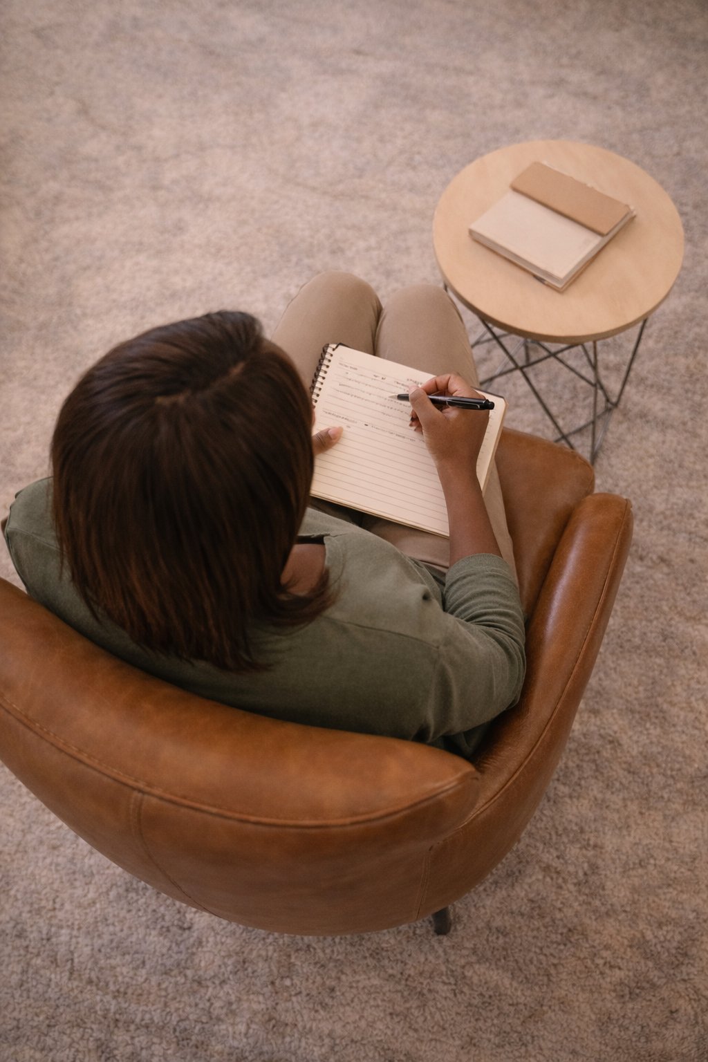 A person with brown hair sitting in a brown leather armchair, writing in a notepad with a pen. A small round table with three closed notebooks is beside them, on a carpeted floor.