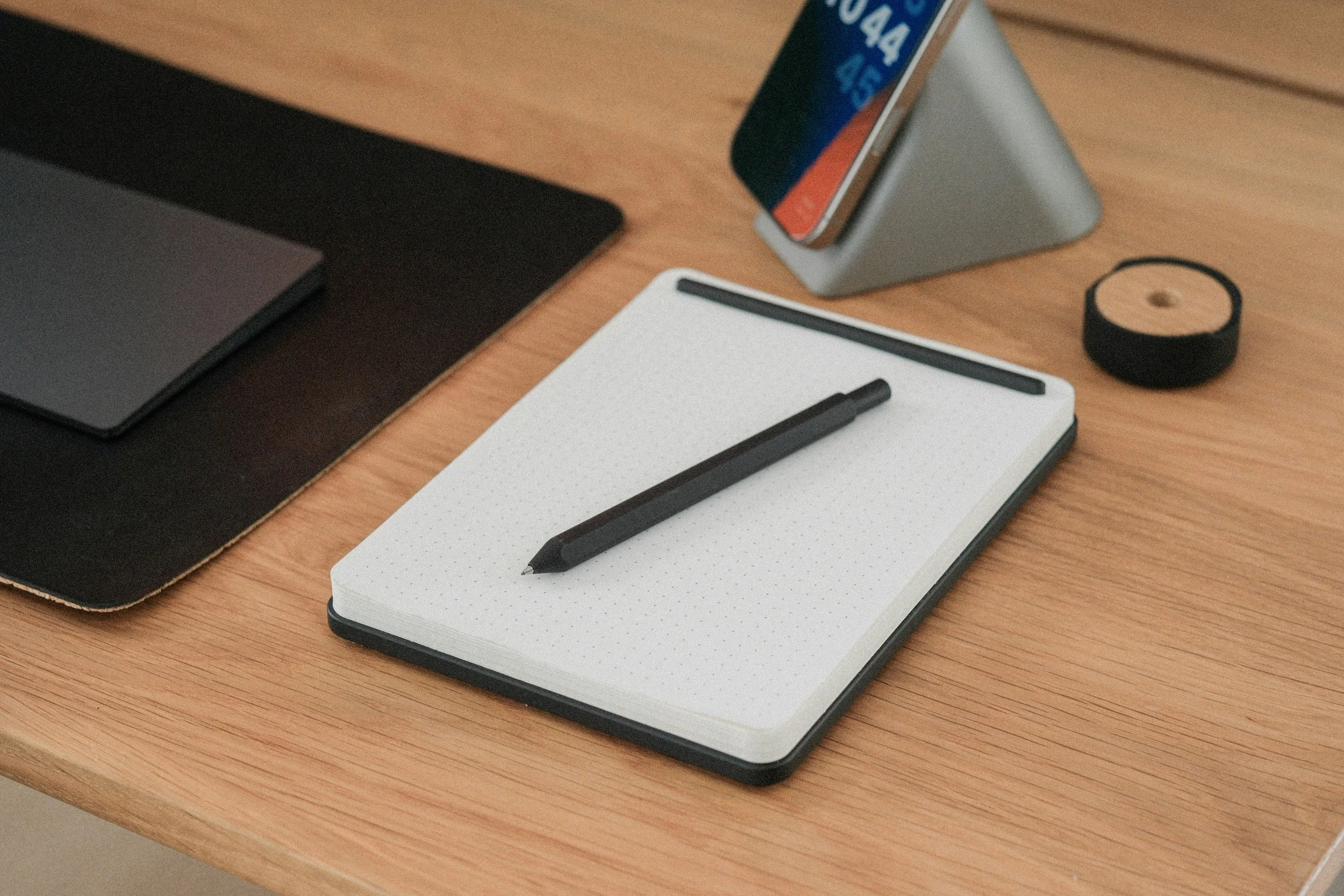 A closed black notebook, a black pen on a dotted white notepad, a smartphone on a silver stand, and a roll of black tape on a wooden desk.