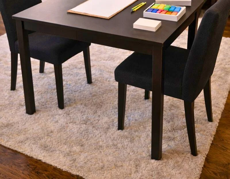 A dark wooden table with two black upholstered chairs, placed on a beige shaggy rug. On the table are colorful chalks, a large sketchpad, and some yellow pens.