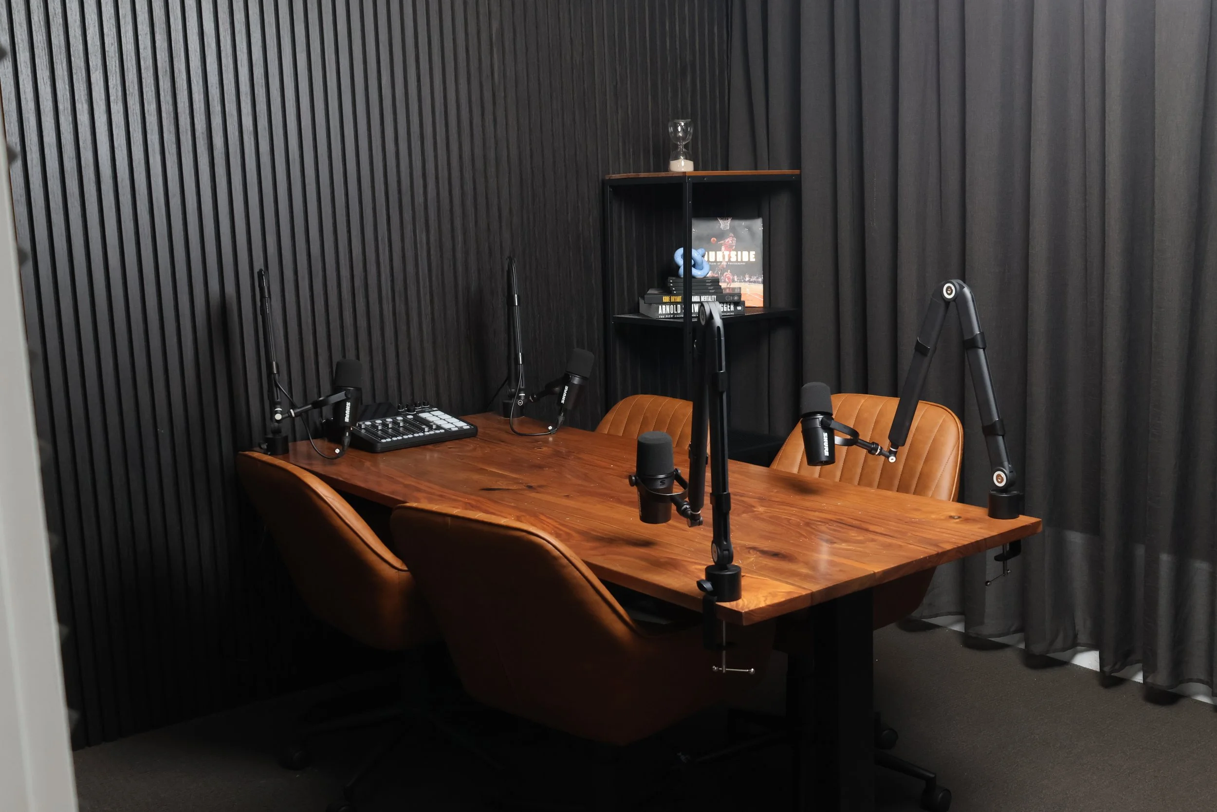 A podcast recording studio with a wooden table, three brown leather chairs, three microphones on adjustable arms, a small audio mixer, and a black bookshelf with books and decorations against a black wall with curtains.