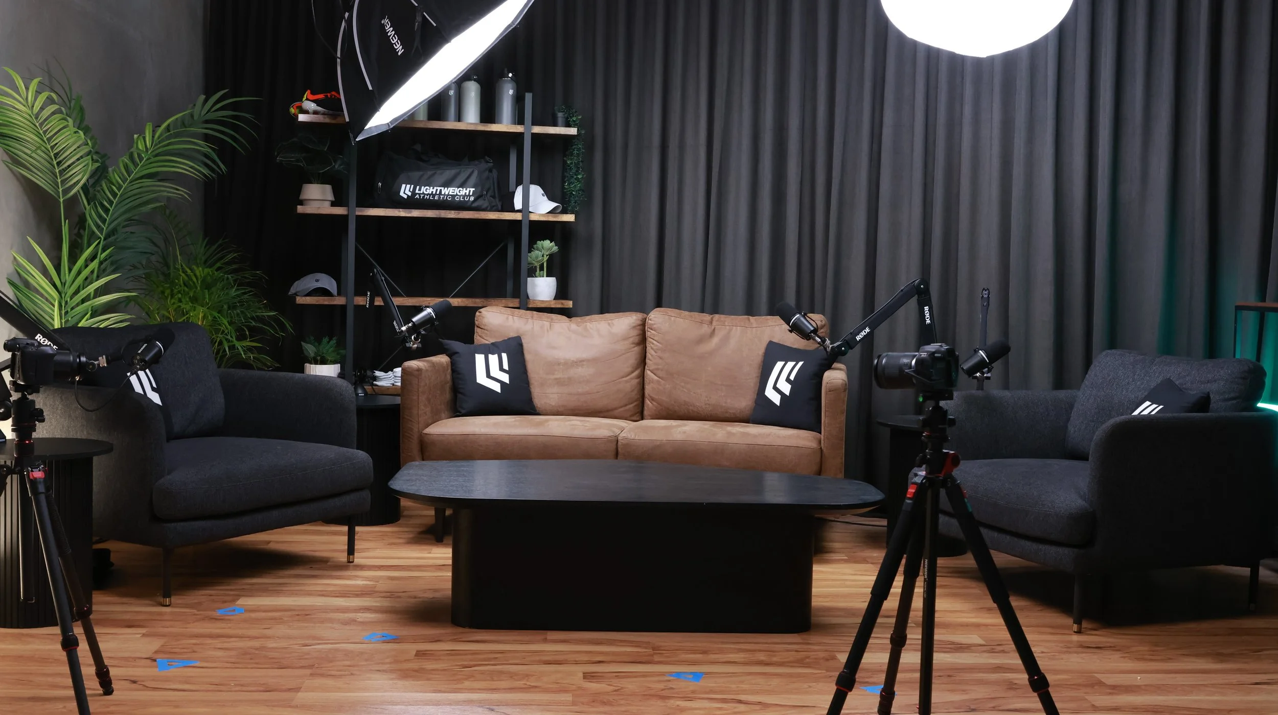 A professional indoor podcast or streaming studio with two black sofas and a tan sofa, black pillows with white logos, a black coffee table, studio lighting, cameras on tripods, green plants, and storage shelves with hats, water bottles, and decorative items, all set against a dark curtain backdrop.