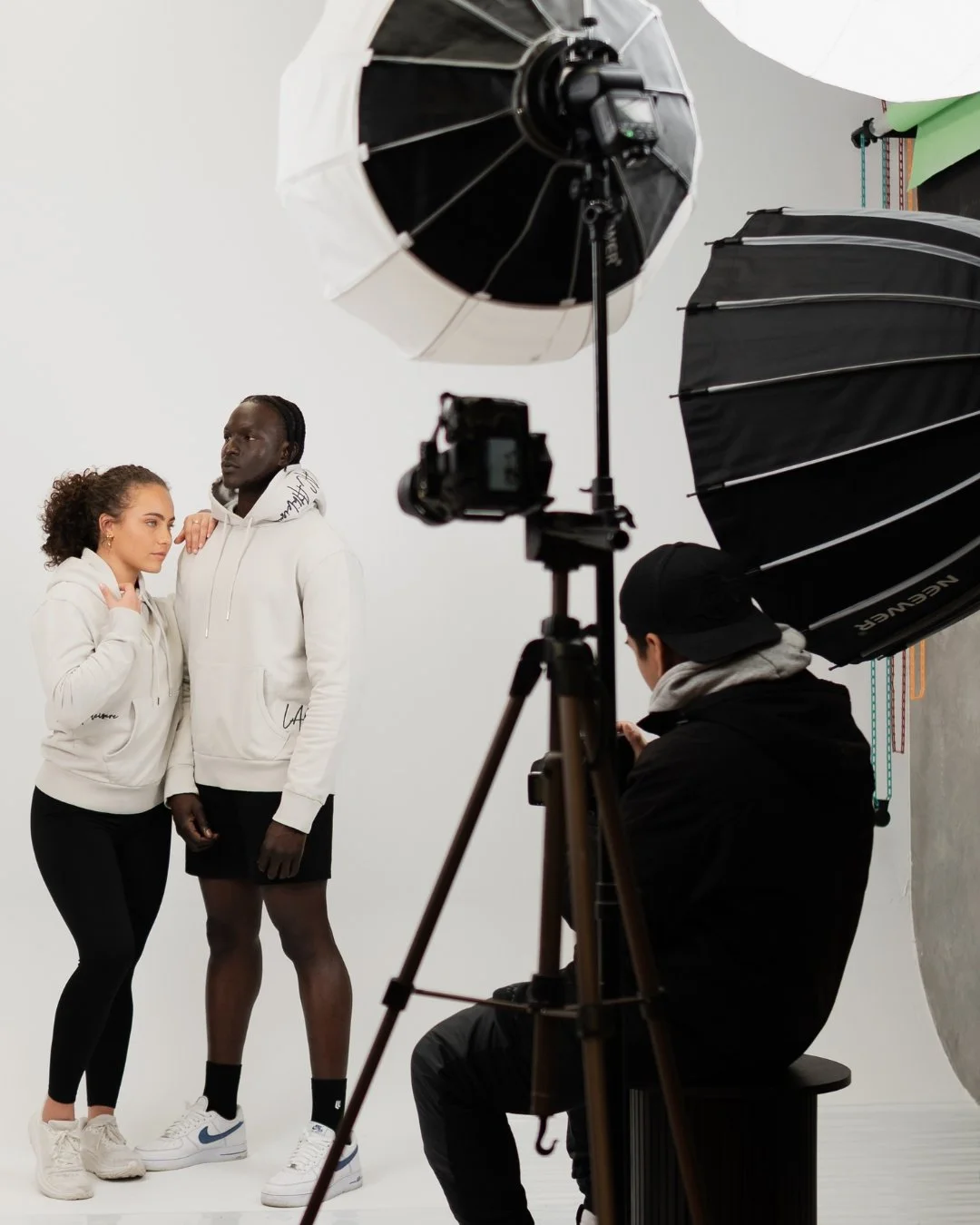 Two young women posing for a photoshoot with professional lighting and camera setup in a studio.