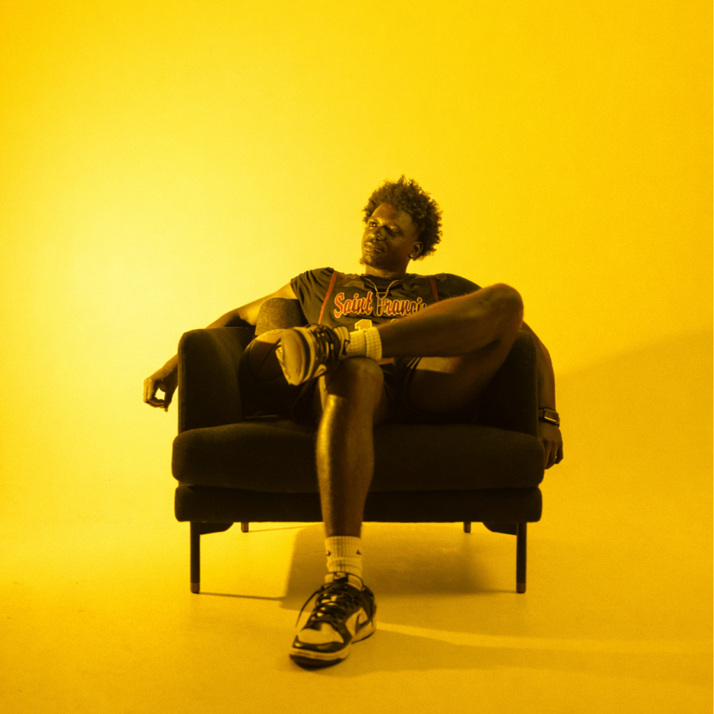 A man sitting on a black armchair against a yellow background, wearing a Saint Francis shirt, shorts, and sneakers, with one leg crossed over the other.